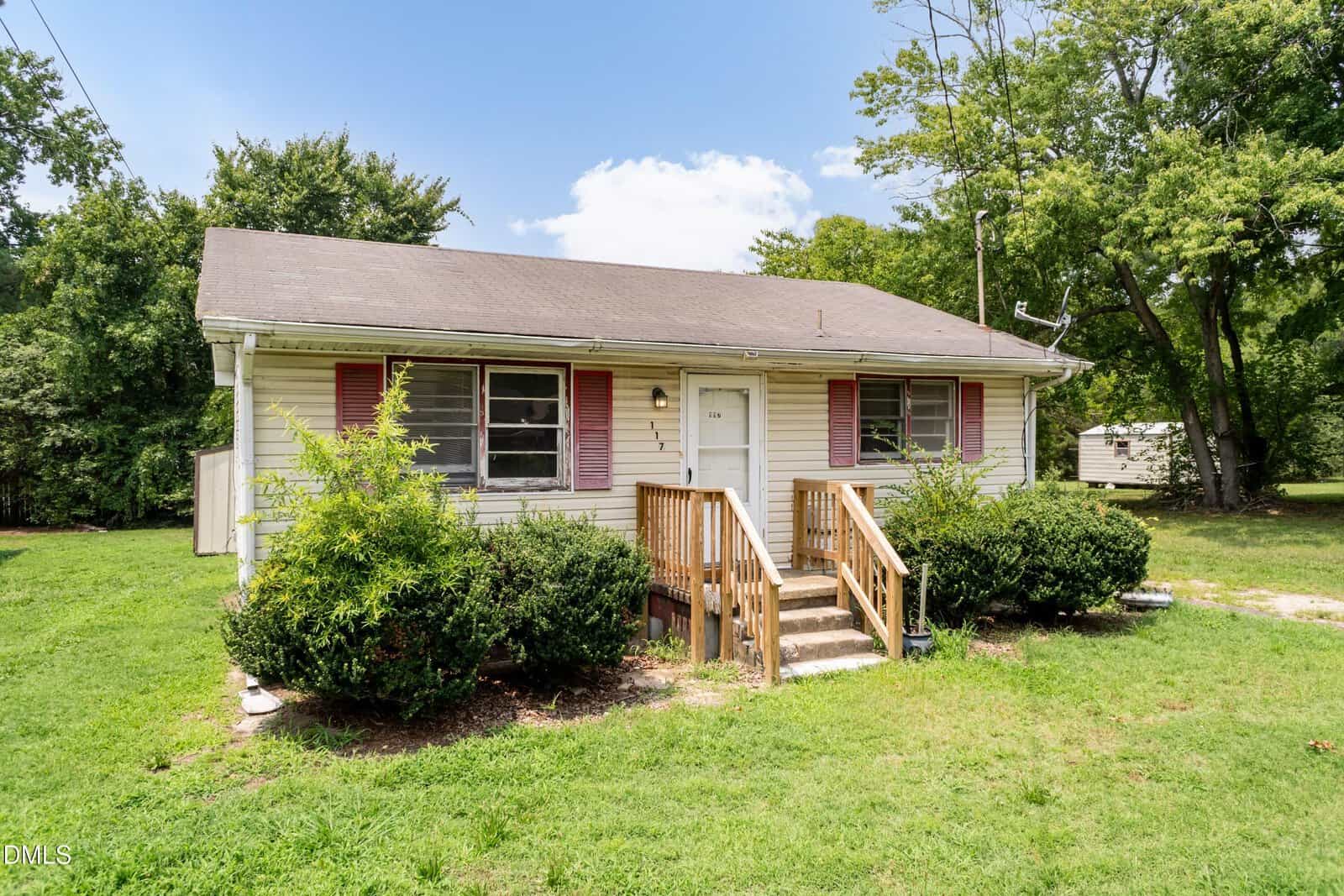 Charming one-story house with beige siding, red shutters, a dark roof, and a small wooden porch with steps located at 117 Warren Avenue in Oxford. Bushes line the front of the house, while a grassy yard and mature trees create a peaceful backdrop under a partly cloudy sky.