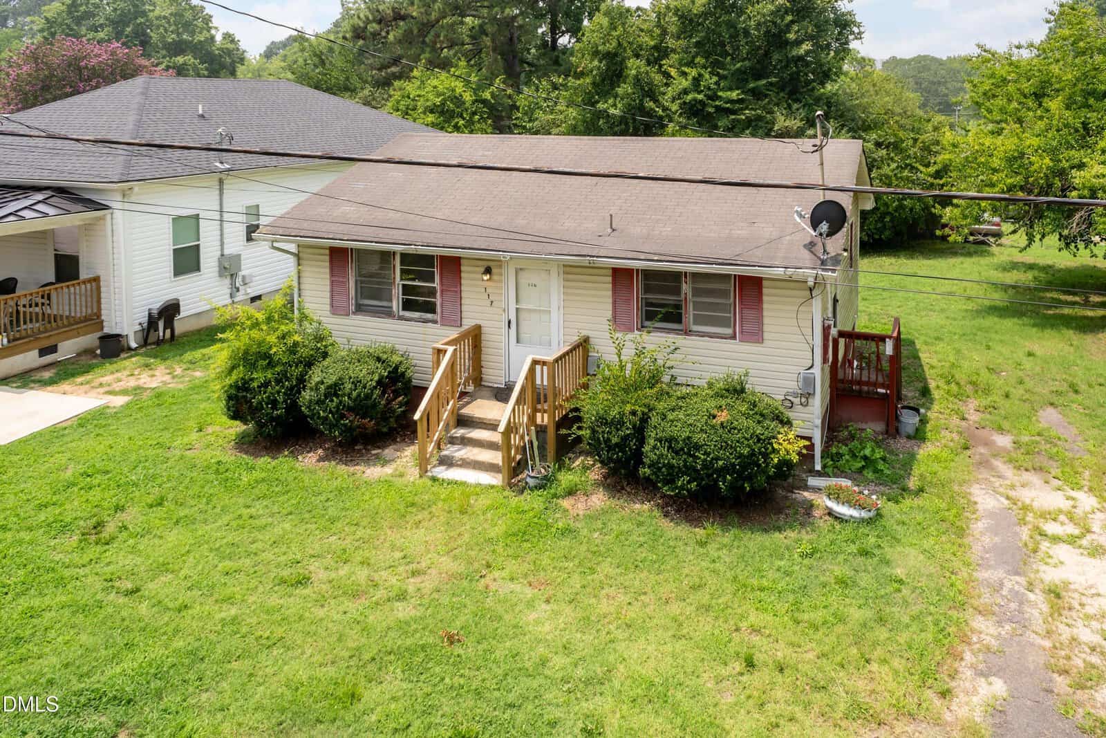 Small single-story house with beige siding, red shutters, and a shingled roof. Front steps lead to a central door. Satellite dish on the roof, bushes in front, and grassy yard with a driveway to the right.
