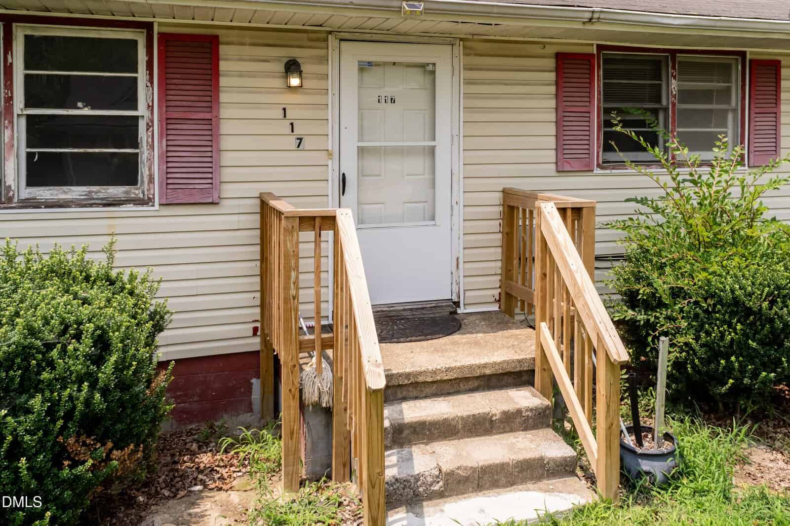 Front entrance of a house with beige siding, red shutters, a white door with a glass storm door, wooden steps and railing, a porch light, house number 117, and bushes on either side of the steps.