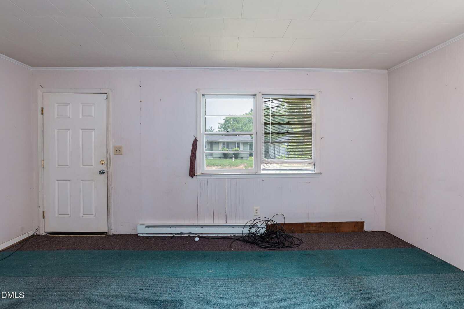 Empty room with teal carpet, white walls, a white door, and two windows. Wires and a wood plank are on the floor below the windows, and blinds are partially open.