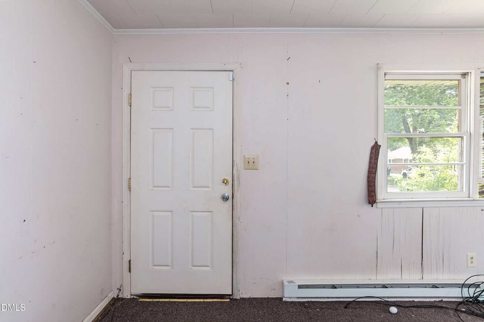 A plain white door with a silver knob is set in a white wall next to a window. The wall and floor appear worn, and there are scuff marks and exposed cables near the baseboard. Green trees are visible outside the window.