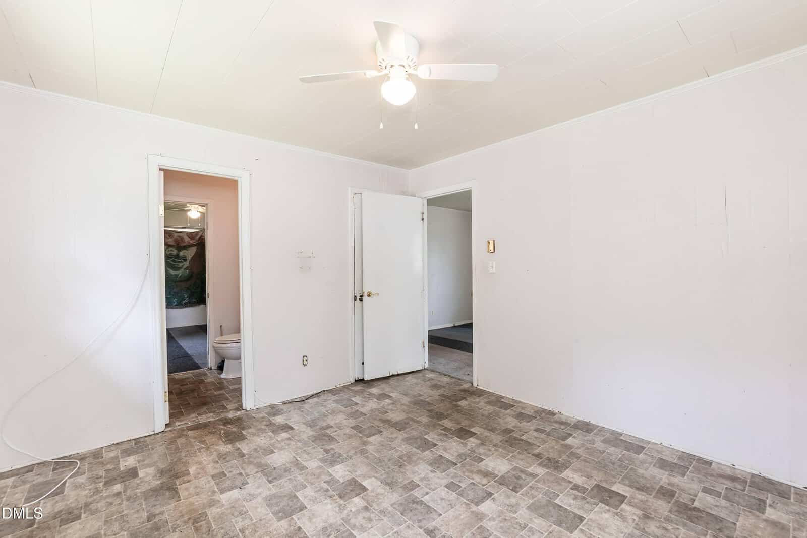 Empty white room with tile flooring, ceiling fan, and two open doors—one leading to a bathroom with a toilet visible, and the other leading to a carpeted room. Walls and ceiling are plain and undecorated.
