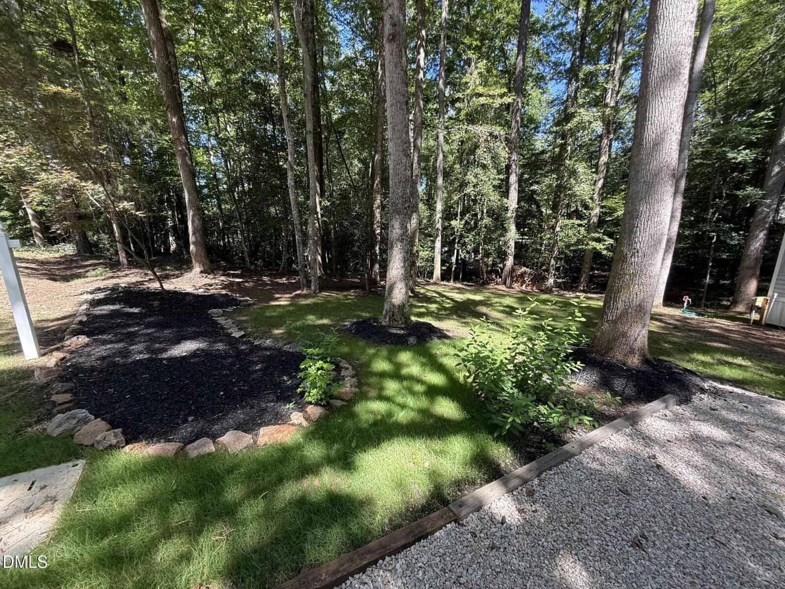 A backyard with tall trees, mulched flower beds edged with stone, green grass, and a gravel area in the foreground, all bordered by a wooded area in the background. Sunlight casts shadows from the trees.