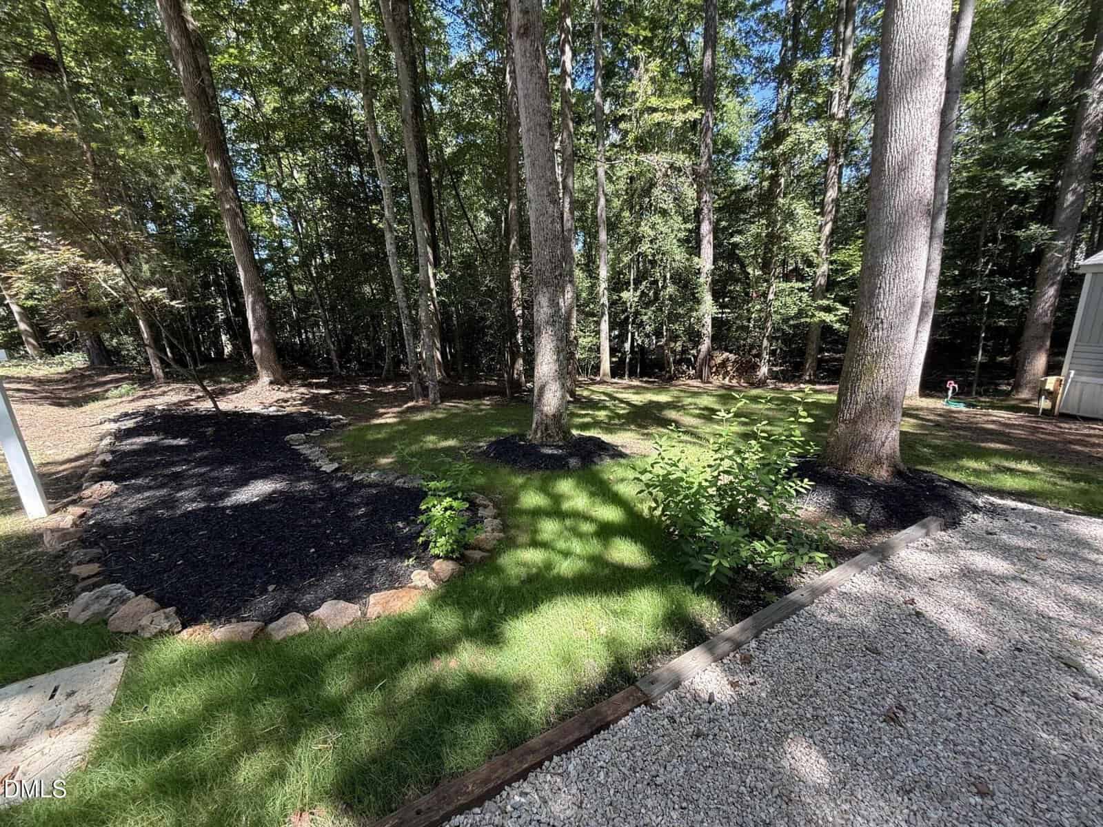 A backyard with tall trees, shaded grass, garden beds bordered by rocks and mulch, and a gravel path beside a wooden deck or patio. Sunlight filters through the trees.