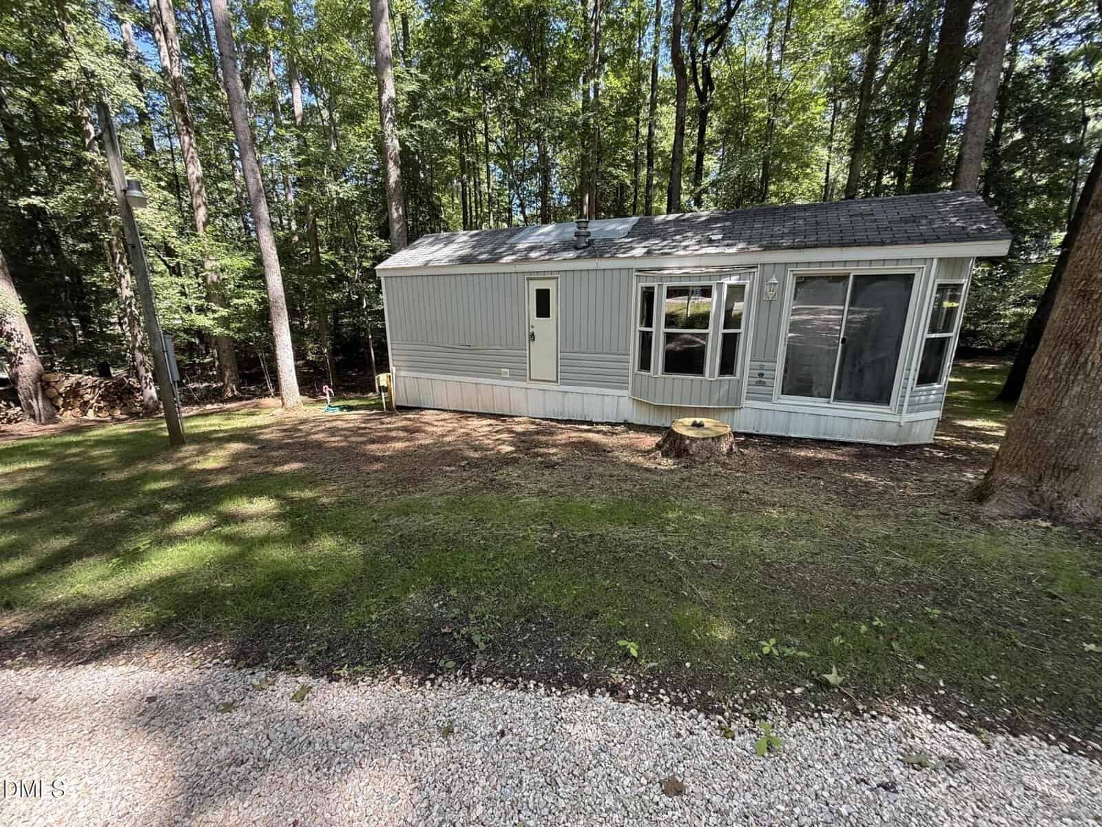 A small, light gray mobile home with a bay window sits on a grassy lot surrounded by tall trees. Sunlight filters through the leaves, and there is a gravel driveway in the foreground.