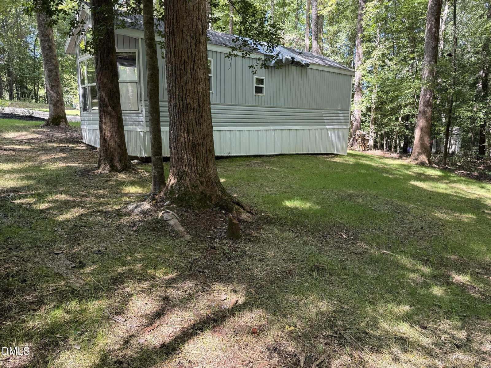A light blue mobile home sits among tall trees on a grassy, shaded lot. Sunlight filters through the leaves, and the ground is uneven with patches of grass and dirt.