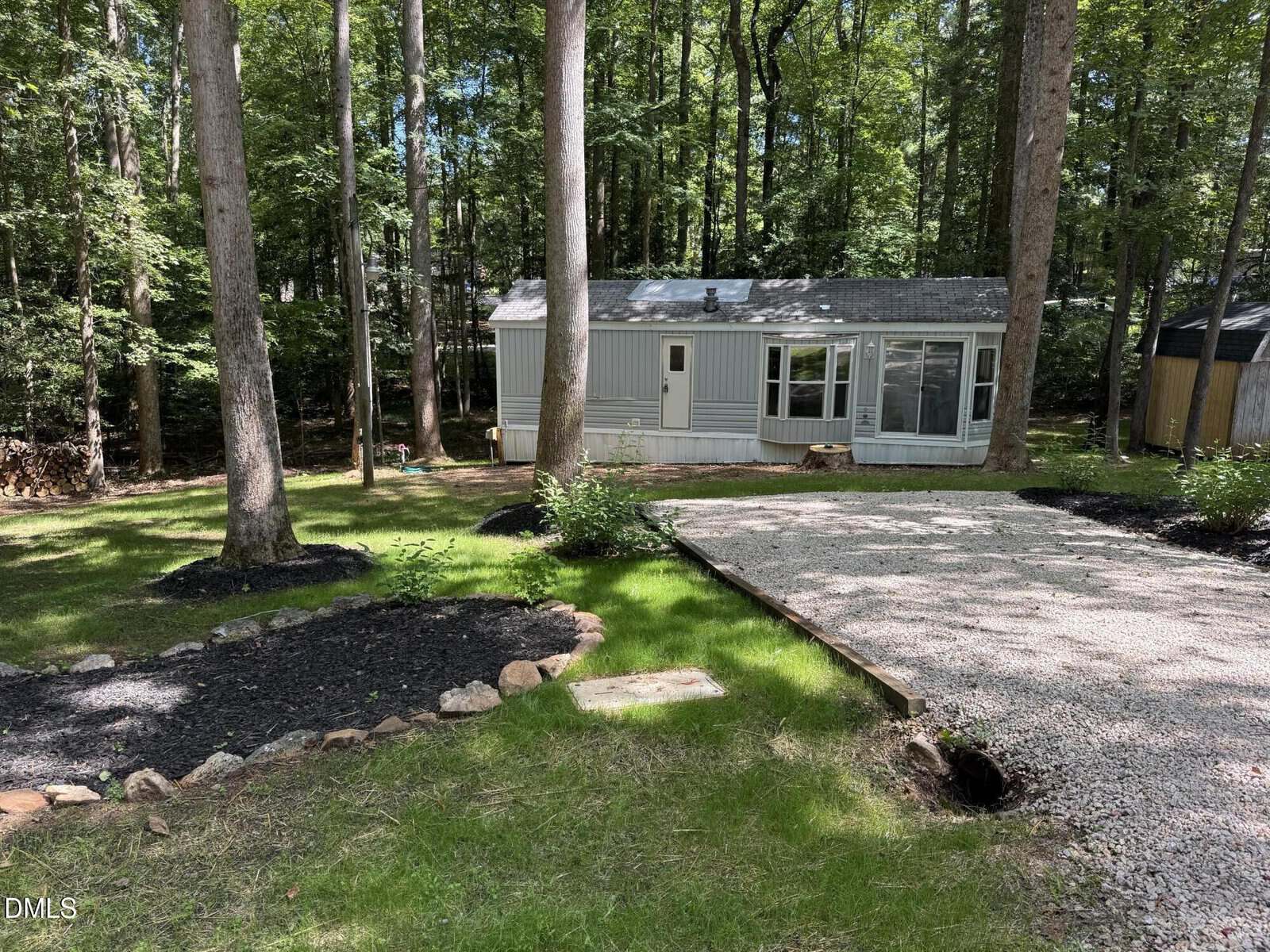 A small gray mobile home sits among tall trees, with a gravel driveway and mulched garden beds in front. The house has a front door, several windows, and is surrounded by green grass and wooded areas.
