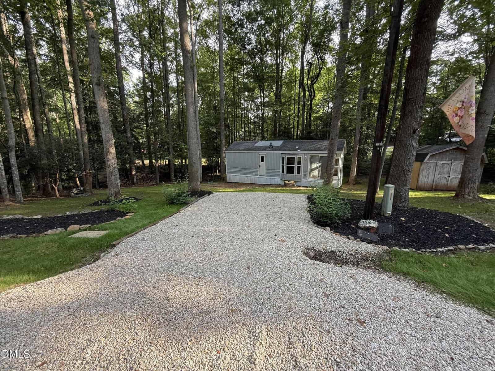A gravel driveway leads to a small, light blue house surrounded by tall trees at 113 Kansas Drive in Louisburg. There are mulched garden beds on each side, a flag on the right, and a shed visible in the background.