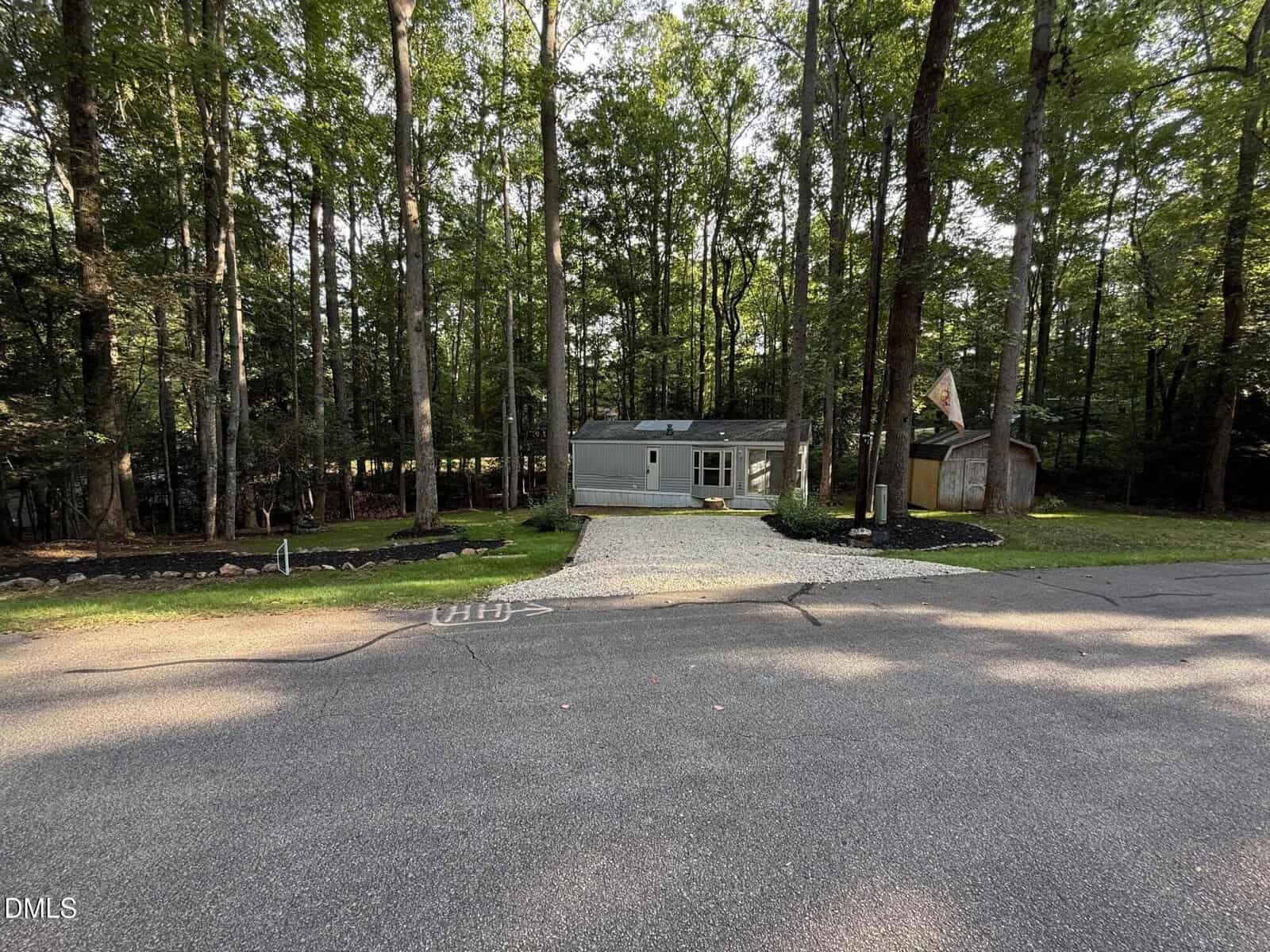A small gray house with a gravel driveway sits among tall trees. A shed is visible on the right, and the yard has simple landscaping with dark mulch and green plants. The scene is viewed from across a paved road.