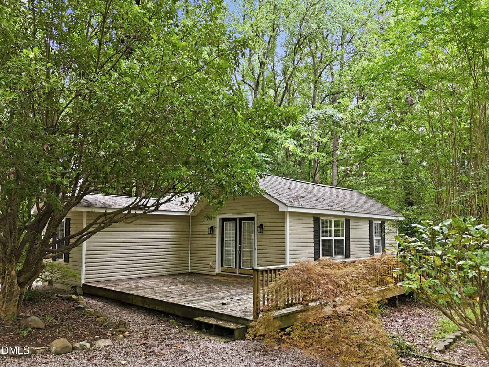 Charming single-story beige house with white trim at 120 Carolina Avenue, Oxford, nestled among dense green trees. The home features a wooden front deck with railings, double glass doors, and several windows. The ground is beautifully landscaped with rocks and shrubs for natural appeal.