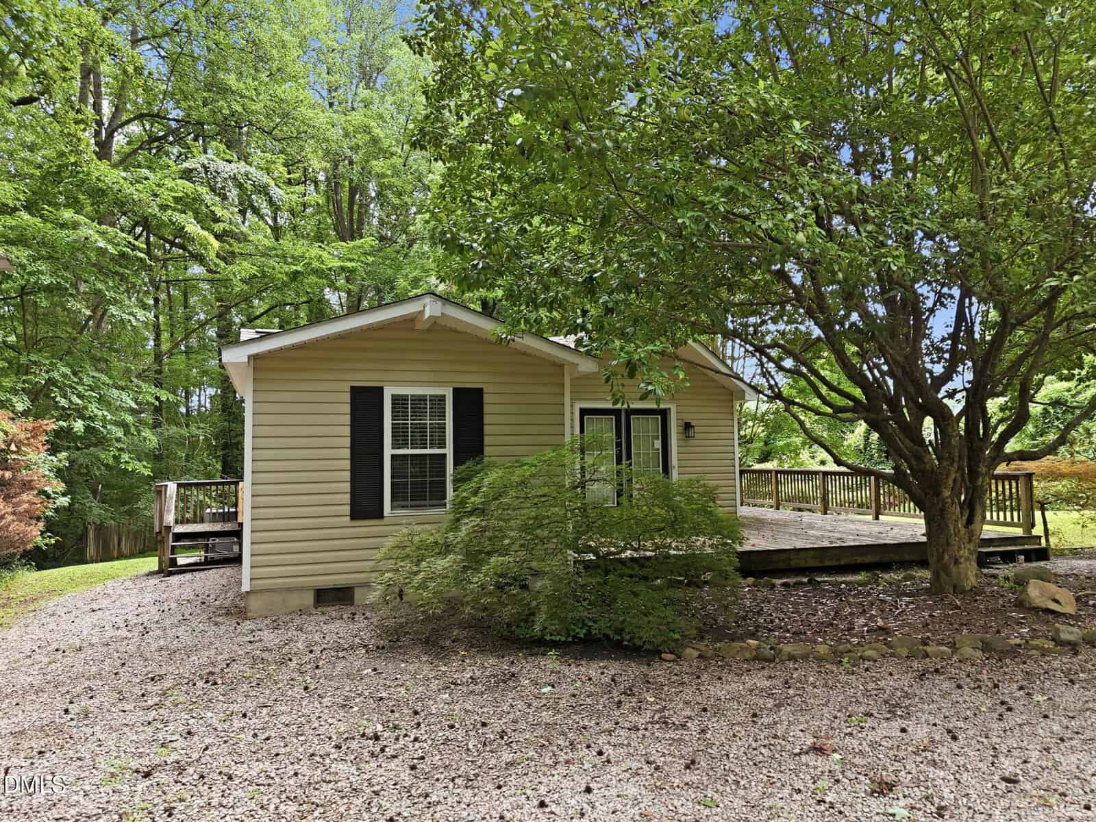 Single-story beige house with black shutters, surrounded by trees. A large tree and green shrub are in the front yard. The house has a gravel driveway and a wooden deck on the right side. The background shows dense green foliage.