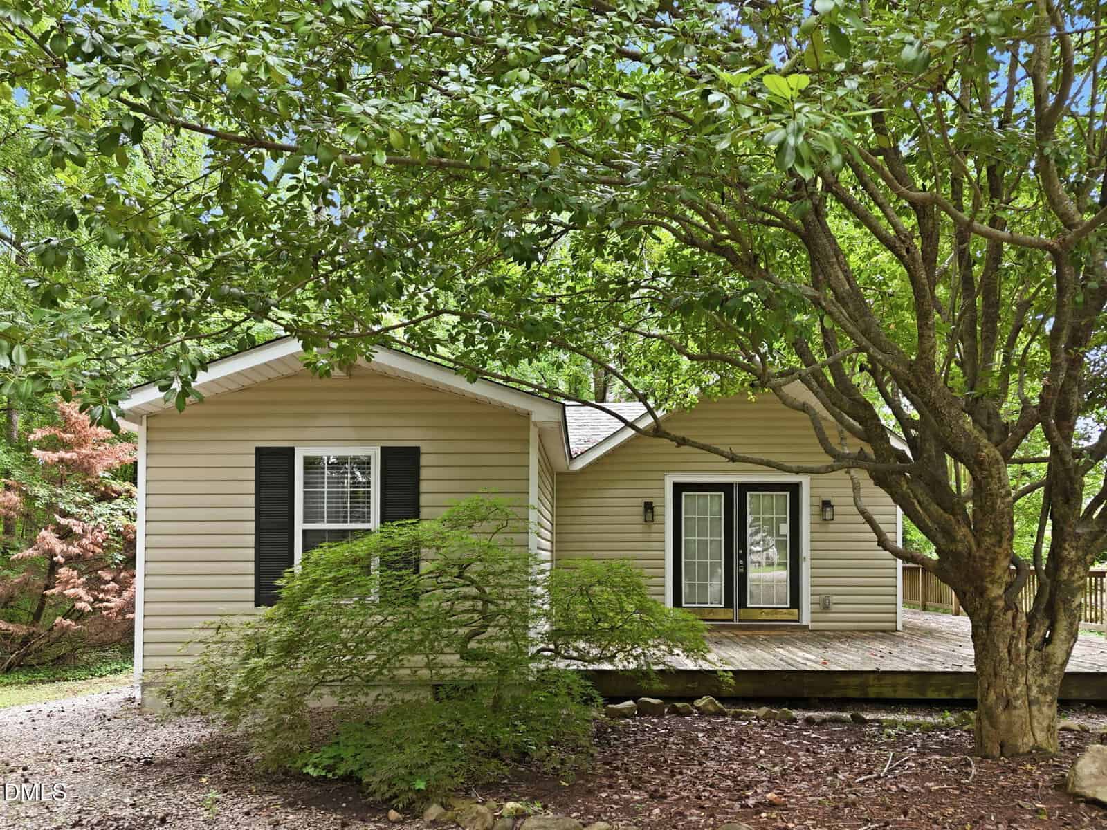 Single-story house with beige siding, black shutters, and large windows. Double glass doors open to a wooden deck. Mature trees and shrubs surround the front yard, providing shade and greenery. Mulch and stones cover the ground.