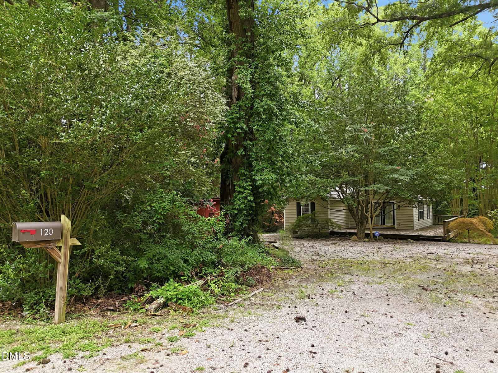 A gravel driveway leads to a light-colored house partially hidden by trees and foliage. A wooden post holds a red mailbox labeled 120 on the left side of the image. The scene is surrounded by greenery.
