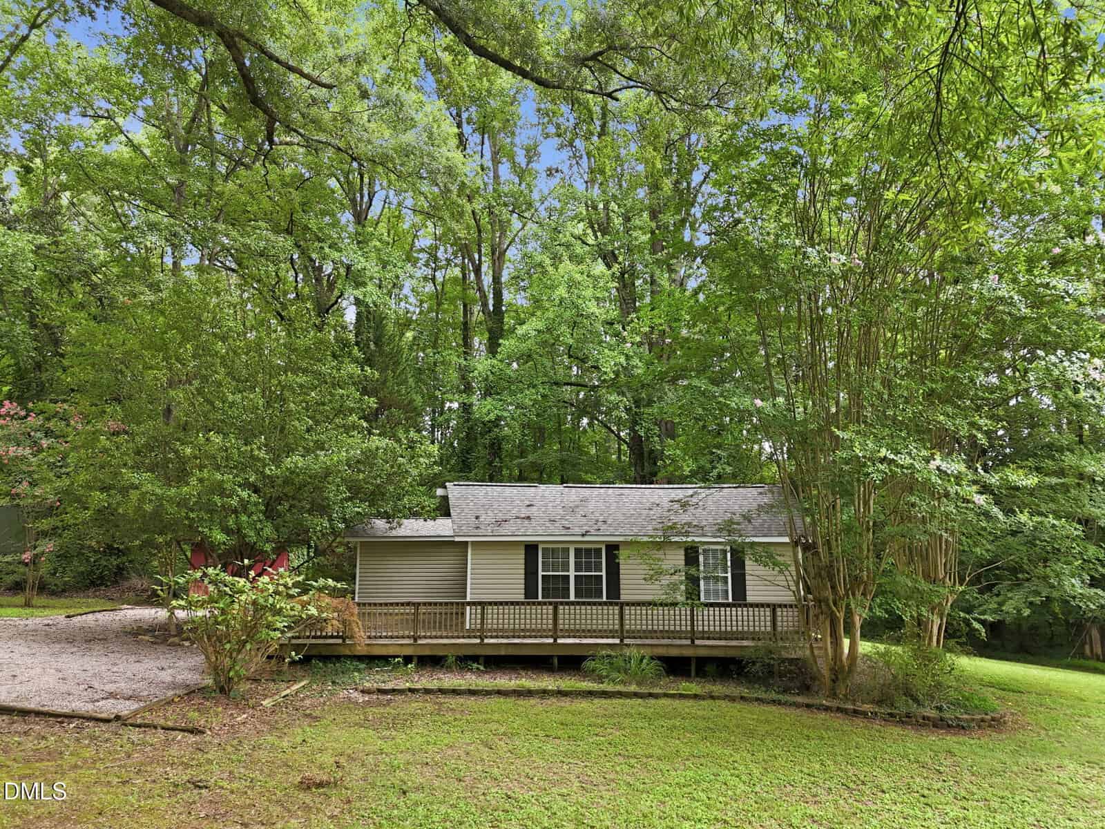 A small, light-colored house with a front porch sits among tall trees and lush greenery. A driveway is visible on the left, and the yard is grassy with shrubs and bushes surrounding the home.