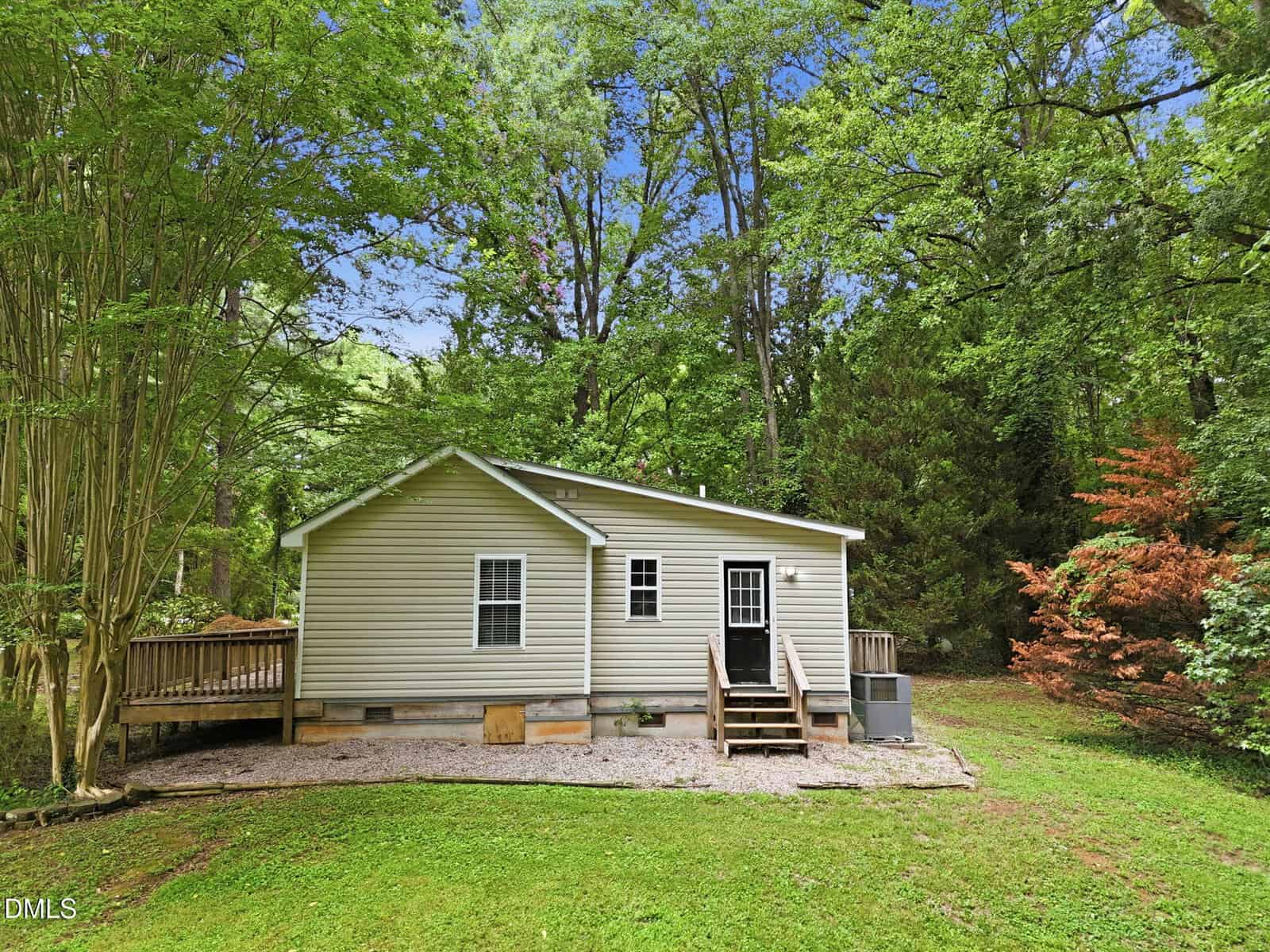 A small beige house with a wooden deck, surrounded by lush green trees and grass. The home has a few steps leading to the back door and is set in a wooded area on a sunny day.
