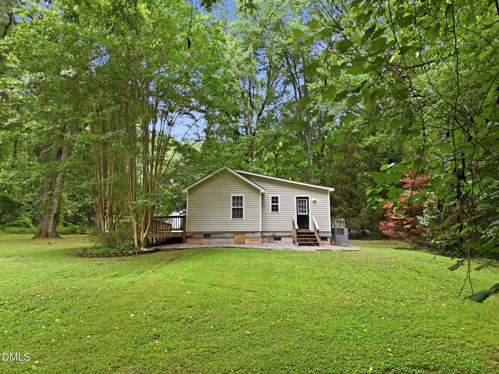 A small beige house with a porch sits in the middle of a grassy yard, surrounded by tall green trees and leafy vegetation under a bright blue sky.