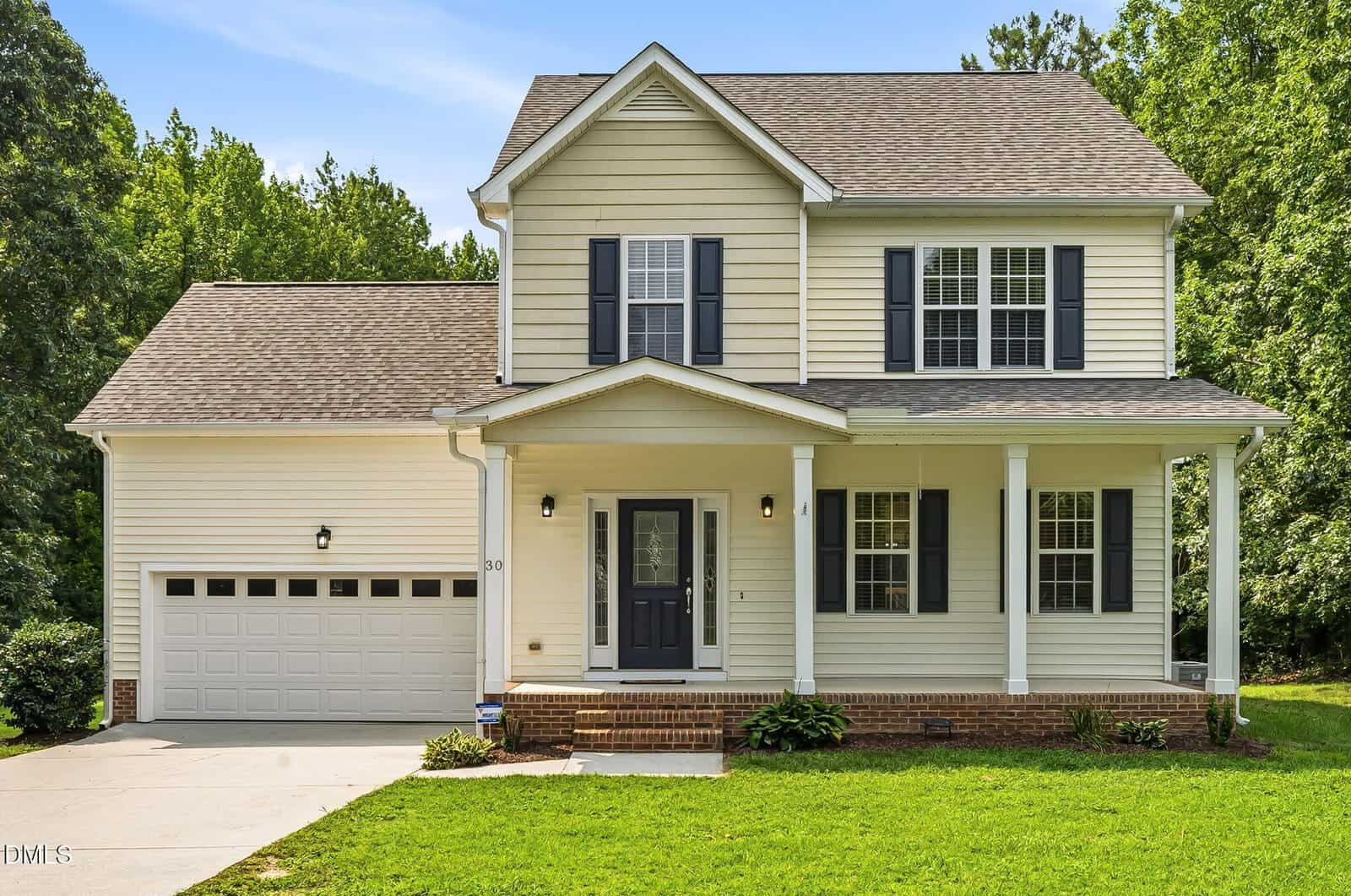 Two-story suburban house with cream-colored siding, dark shutters, a covered front porch supported by columns, and an attached two-car garage. Located at 30 Fern Court in Louisburg, the home features a lush green lawn and is surrounded by trees under a clear blue sky.