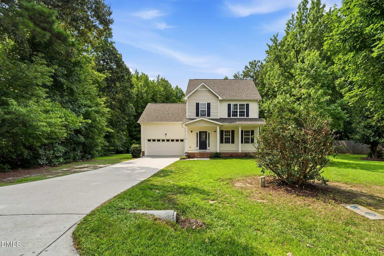 Two-story beige house with a covered front porch, attached two-car garage, and black shutters. Concrete driveway leads up to the house, surrounded by a large green lawn and bordered by trees.