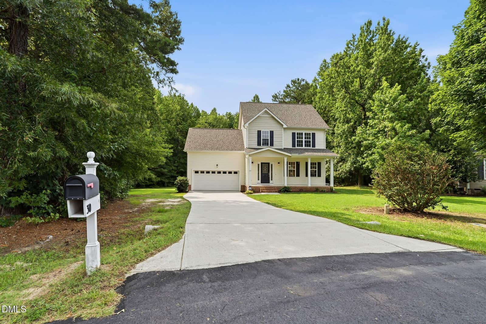 A two-story beige house with dark shutters, a front porch, and an attached garage sits at the end of a long driveway, surrounded by a green lawn and trees. A white mailbox is at the driveway’s entrance.