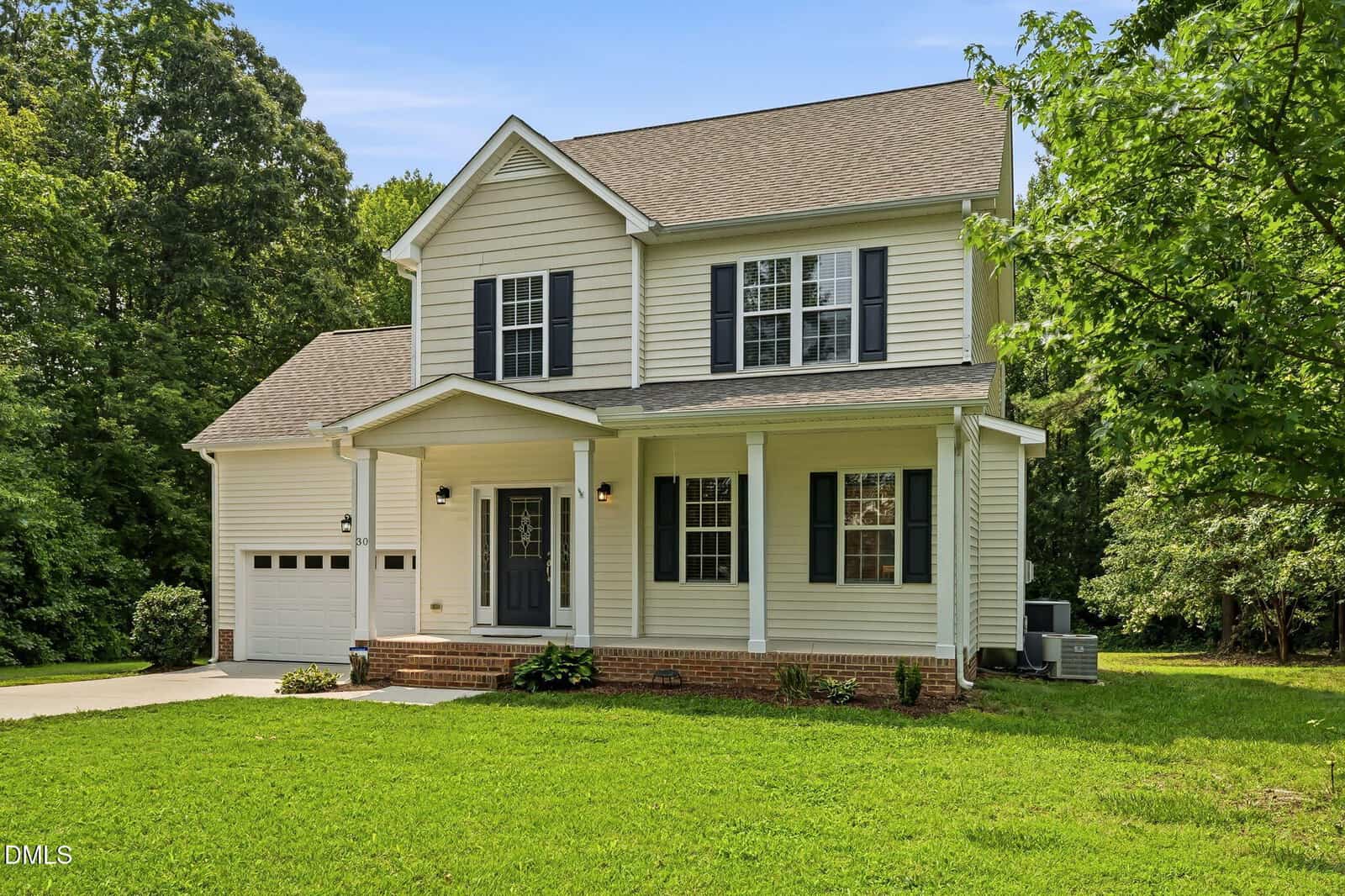 A two-story beige house with a covered front porch, black shutters, and an attached garage, surrounded by green grass and trees under a clear blue sky.