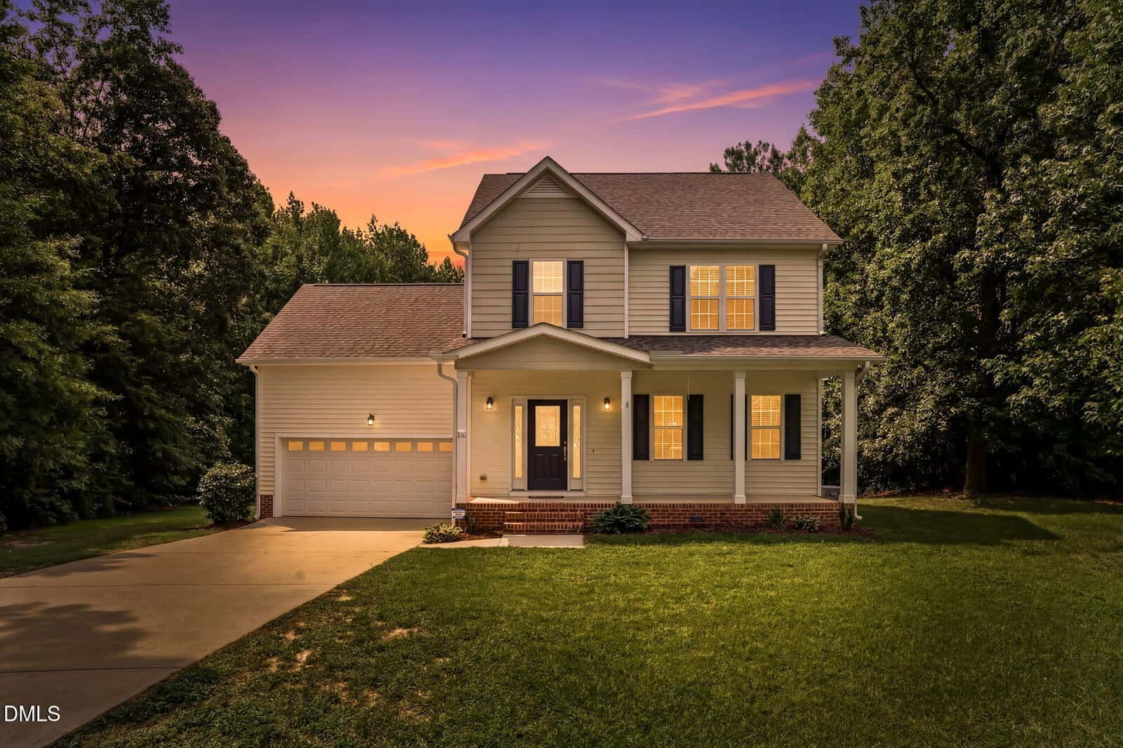 A two-story house with gray siding, a front porch, and an attached two-car garage is shown at sunset. The house is surrounded by grass and trees, with an illuminated sky in the background.