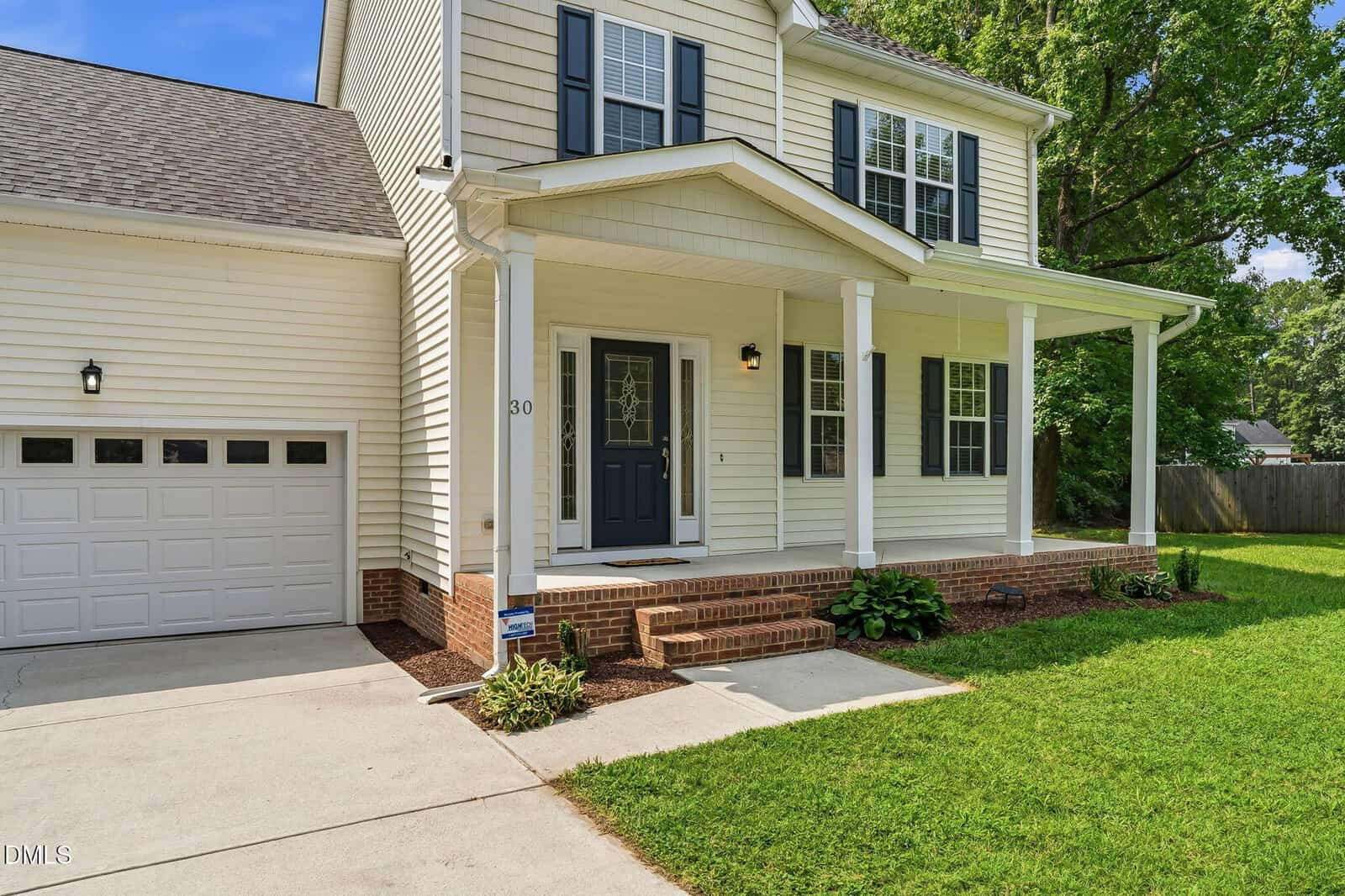 Two-story beige house with black shutters, white columns, a front porch, attached garage, green lawn, and bushes along the brick steps leading to the front door.