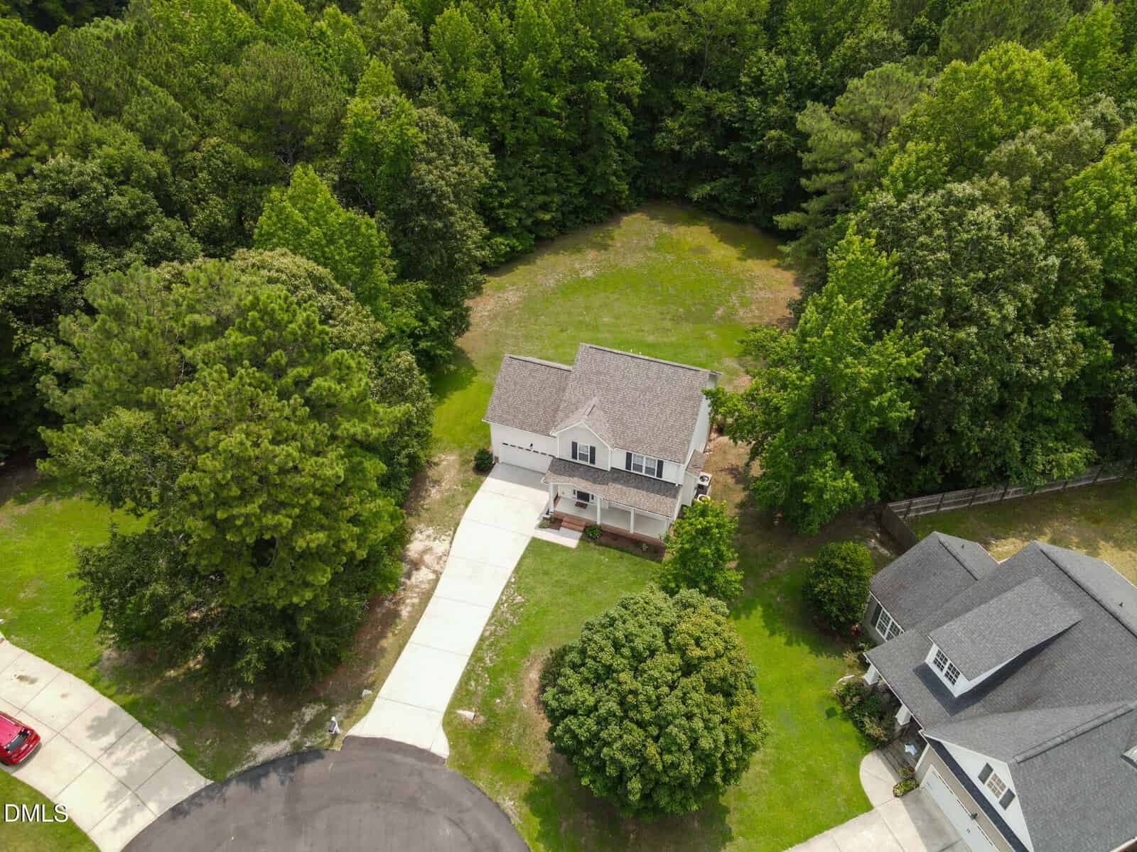 A two-story house with a driveway is located at the end of a cul-de-sac, surrounded by trees and a large grassy yard. The home is adjacent to another house and bordered by a wooded area in the background.