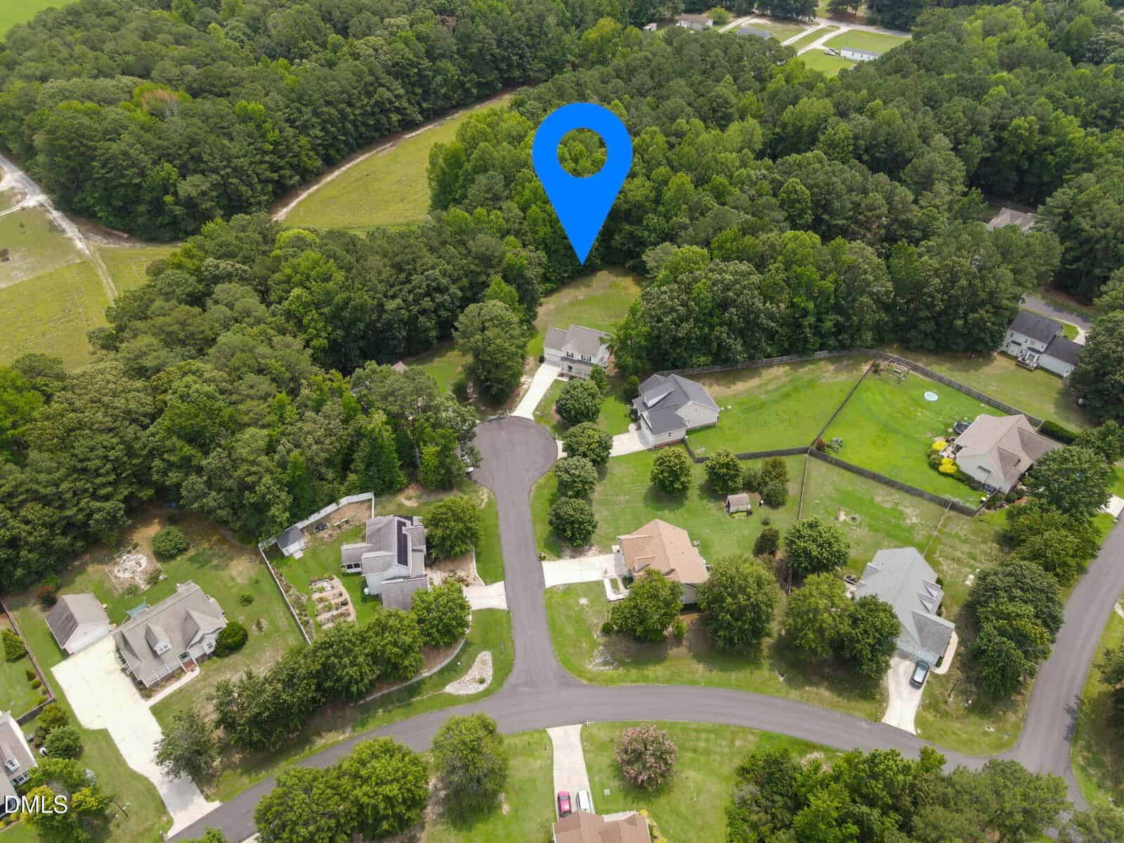 Aerial view of a suburban neighborhood with several houses, curved streets, green lawns, and dense surrounding trees. A large blue location marker highlights a specific house near the edge of the wooded area.