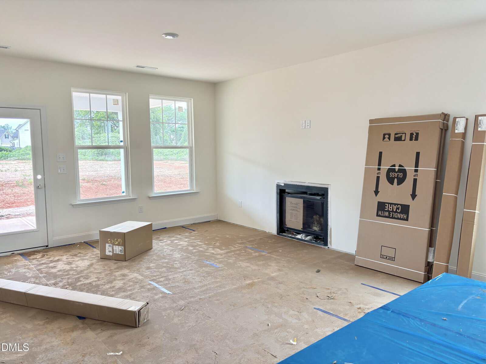 Unfurnished living room with covered floors, a boxed fireplace, and several unpacked cardboard boxes. Two windows and a glass door let in natural light. The walls are painted white.