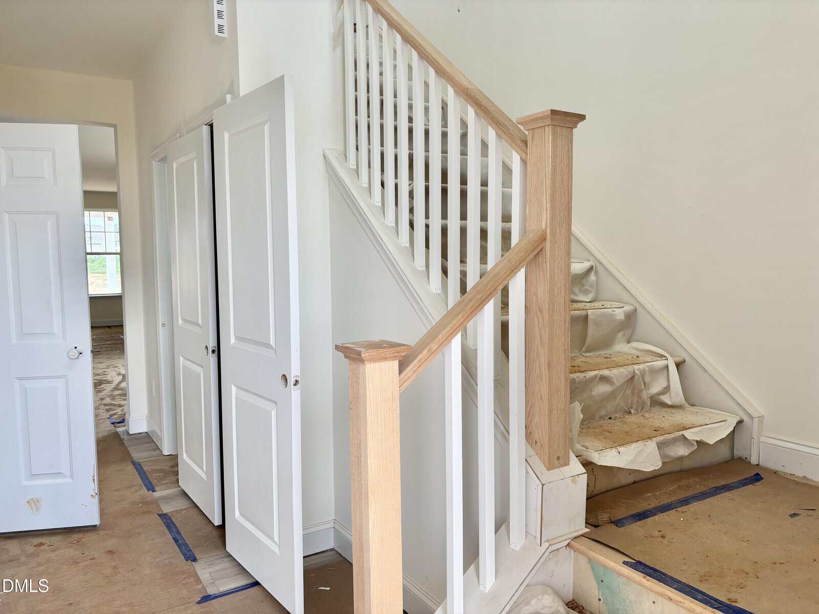 A staircase with light wood handrails and white spindles is next to a hallway with white doors. The stairs are partially covered with protective material, and blue tape lines the floor edges. Walls are painted white.