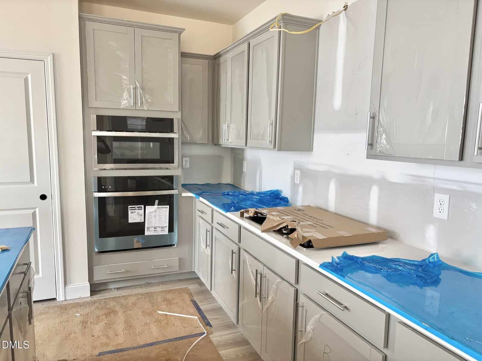 Modern kitchen under construction with gray cabinets, appliances installed, cardboard and blue plastic covering some surfaces, and unfinished walls. Some cabinet doors have protective plastic, and an exposed wire hangs from the ceiling.