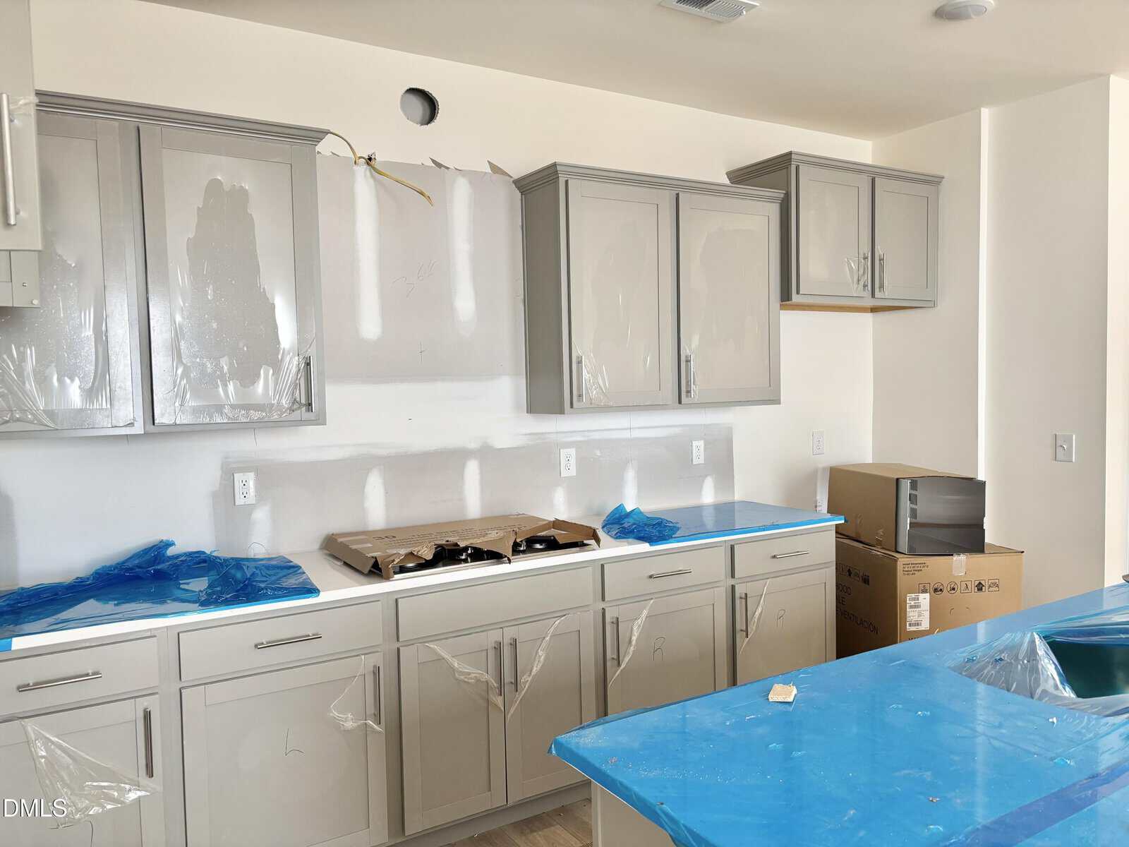 A new kitchen under construction with gray cabinets, blue protective coverings on surfaces, a stove area without appliances installed, and cardboard boxes on the floor and countertop. Plastic sheets partially cover the cabinet doors.