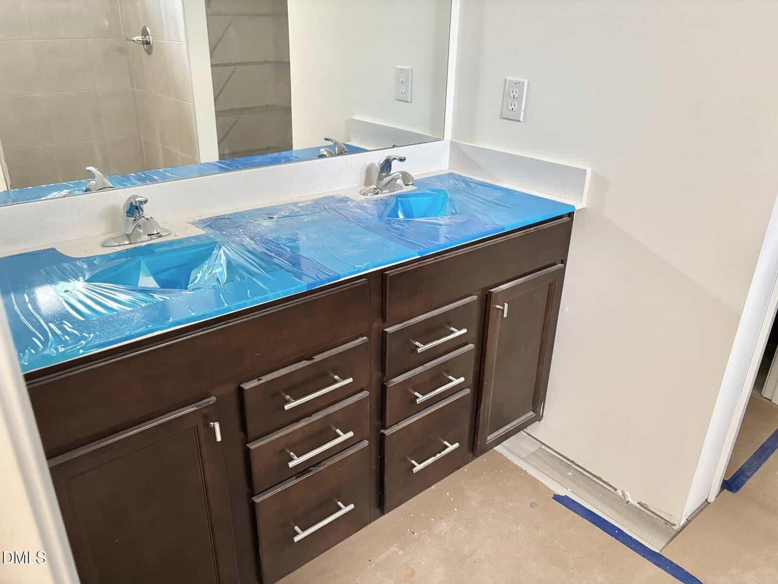 Bathroom vanity with dark wood cabinets and silver handles, double sinks covered with blue protective plastic, and a countertop under a wall mirror. The room appears to be under construction or recently installed.