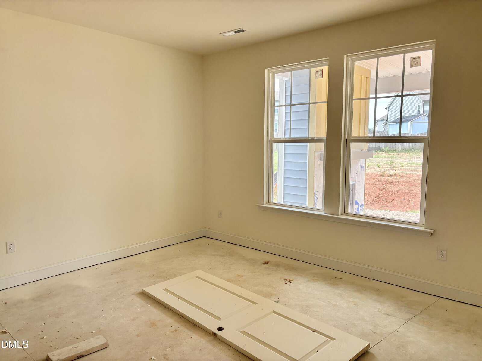 Empty room with cream-colored walls, two large windows, and an interior door lying on the unfinished floor. Daylight comes through the windows, revealing the view of a neighboring house and bare ground outside.