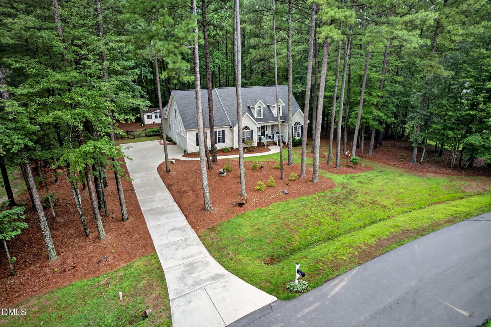 A beige house with a gray roof sits among tall pine trees. A long driveway curves from the road to the front entrance. The lawn is bordered by mulch, and a mailbox is visible at the edge of the driveway near the street.
