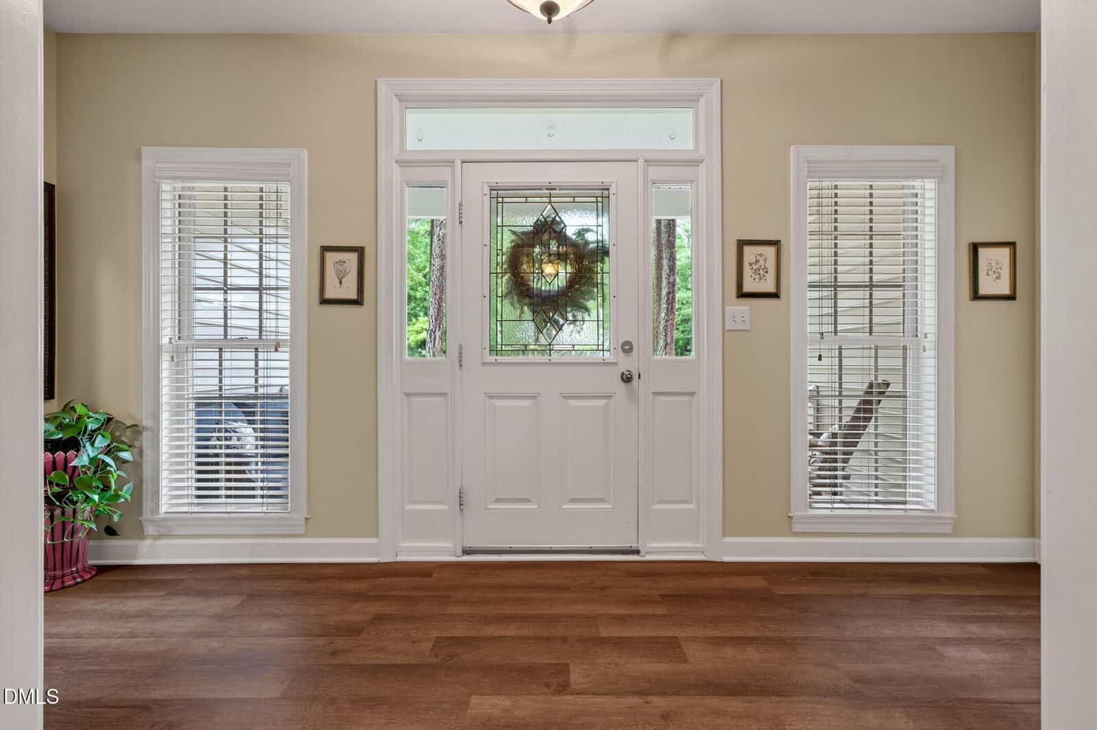 Bright entryway with a white front door, two tall windows on each side, light tan walls, wood floor, framed art, and a potted plant in the left corner. A wreath hangs on the glass of the front door.