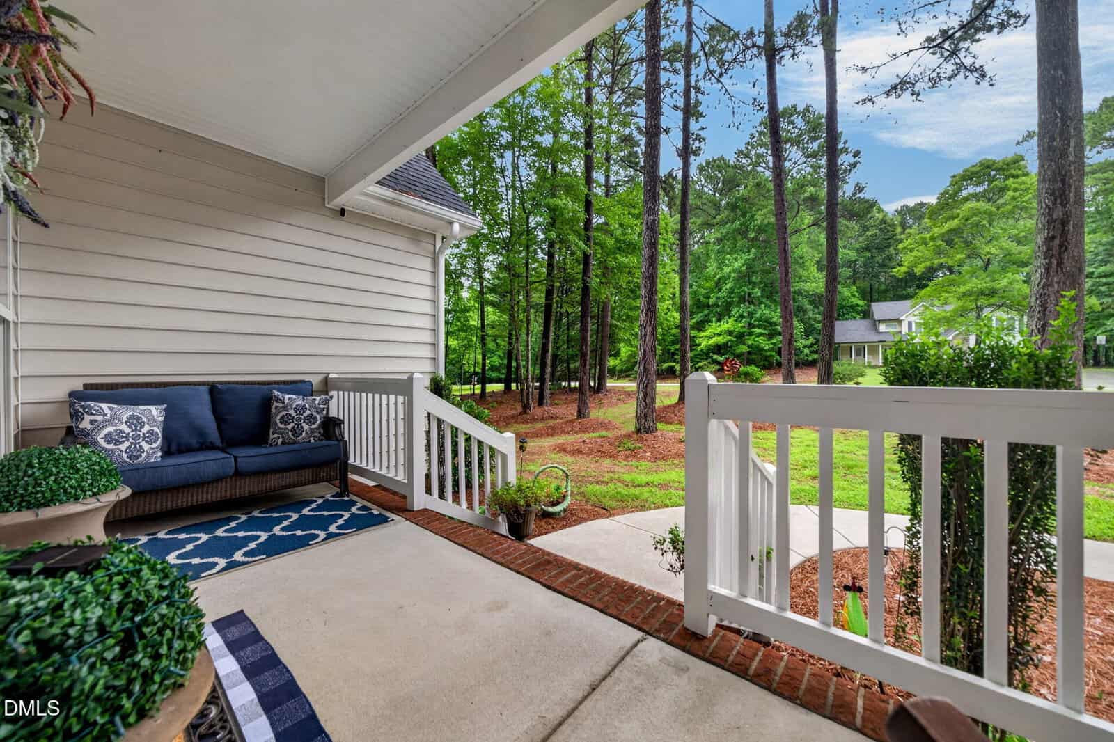 A cozy porch with a navy outdoor sofa, patterned rug, potted plants, and white railing overlooks a yard with pine trees and a curved driveway leading to houses in a wooded neighborhood.