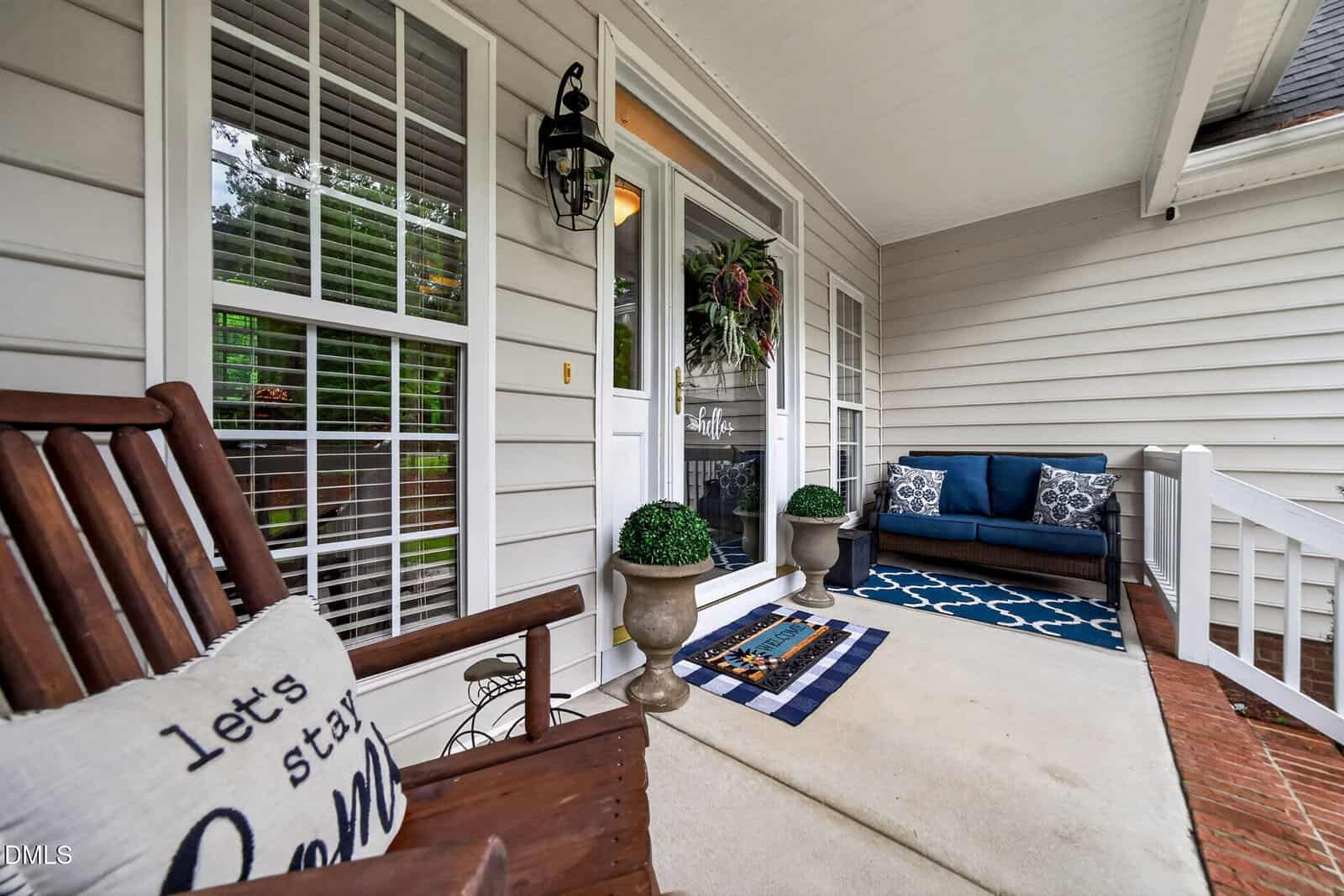 Cozy front porch with a wooden bench, pillow reading “let’s stay home,” potted plants, blue cushioned loveseat, patterned rug, welcome mat, and white siding, creating an inviting, homey atmosphere.