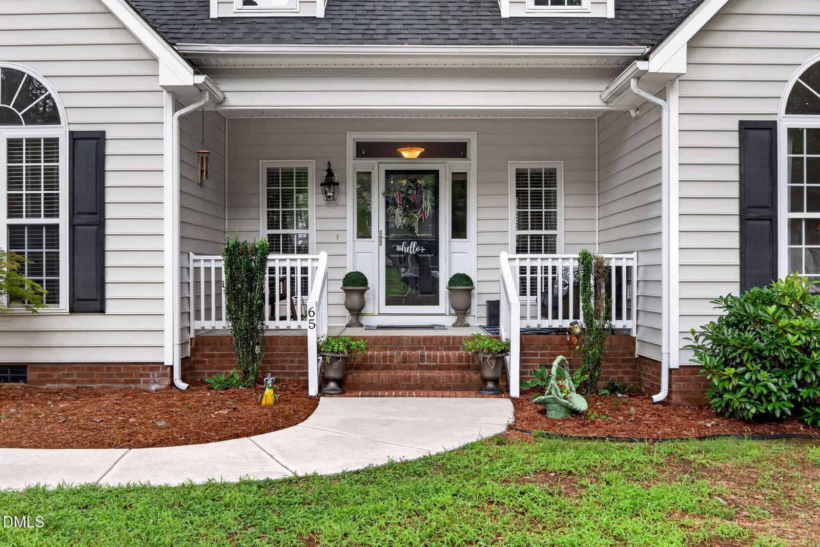 A front porch with white railings, brick steps, two potted plants, a welcome sign on the door, and decorative items on either side. The house exterior is light-colored with black shutters and mulch landscaping.