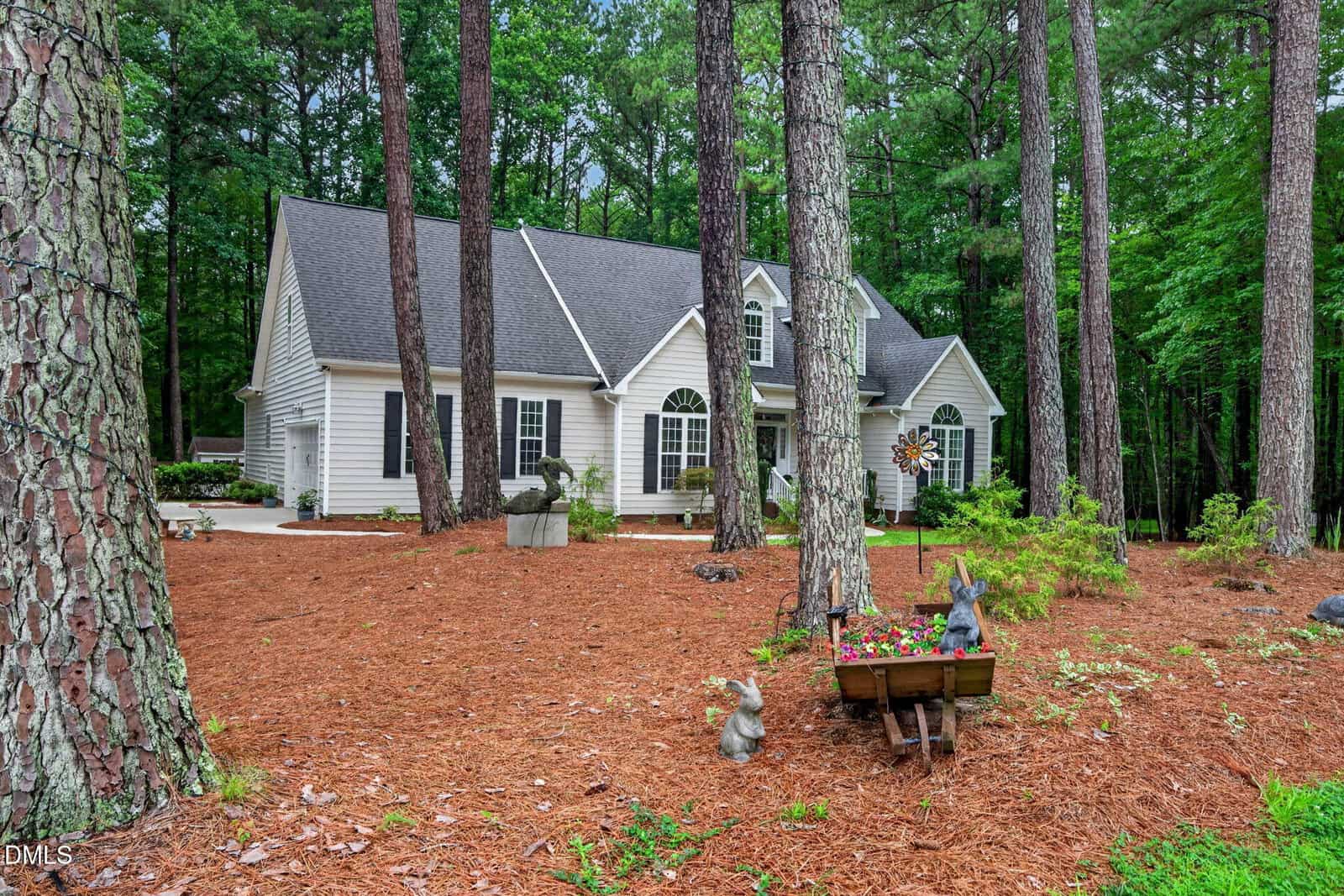 A white house with dark roof sits among tall pine trees. The yard has pine straw ground cover, small shrubs, and lawn ornaments, including a wheelbarrow planter with flowers and animal statues. The background is wooded.