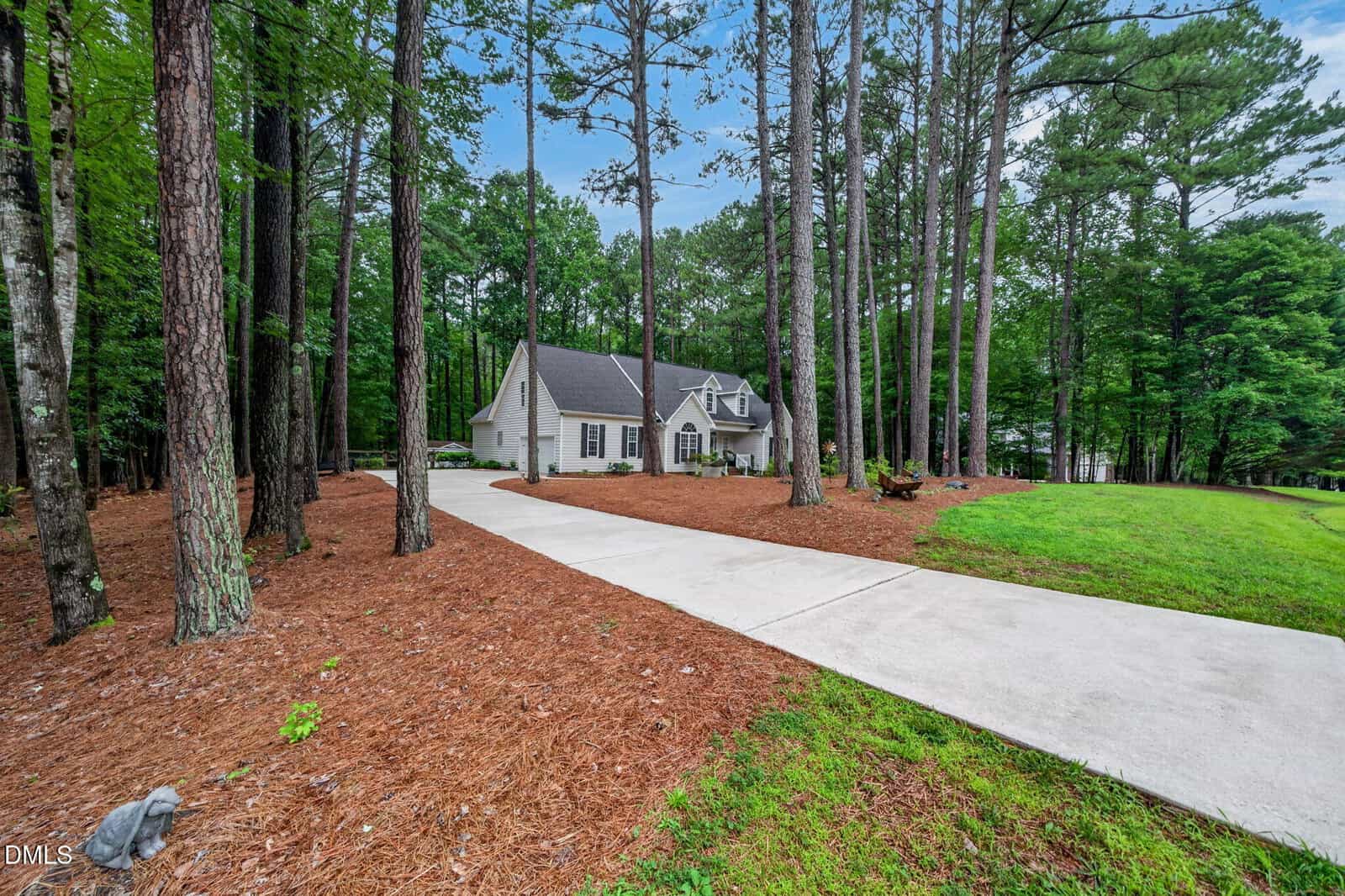 A white two-story house with dormer windows sits at the end of a long concrete driveway at 65 Summit Ridge Court in Youngsville, surrounded by tall pine trees and mulch, with a green lawn on one side under a blue sky with clouds.