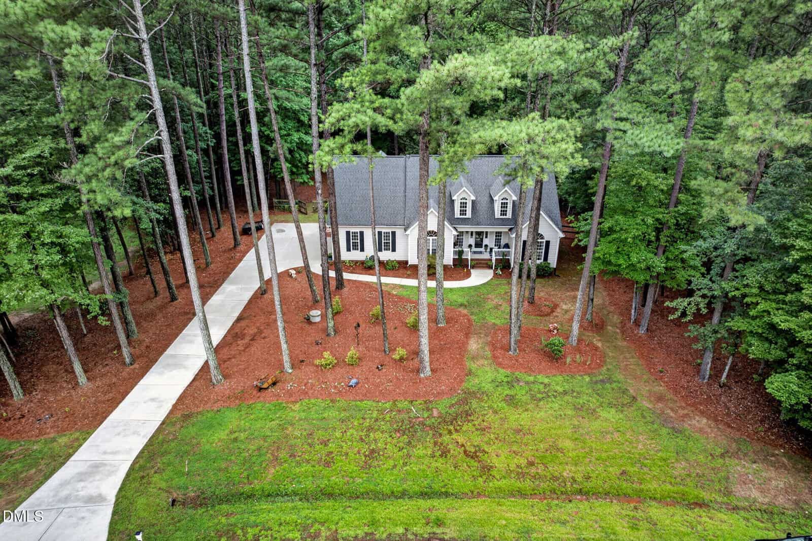 A two-story house sits among tall pine trees, with a curved driveway leading to a detached garage. The yard is landscaped with mulch beds and grass, surrounded by a dense wooded area.