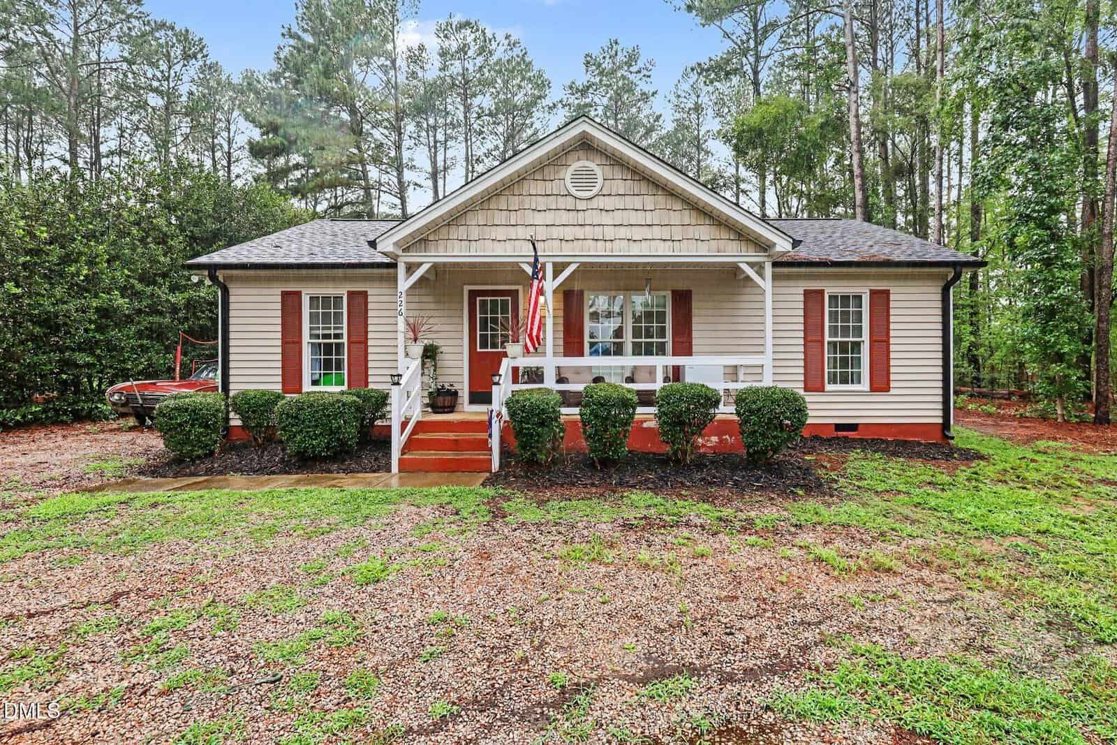 A single-story beige house with a front porch, white trim, and red shutters sits surrounded by trees at 226 Shawnee Drive in Louisburg. Six trimmed bushes line the porch, and a U.S. flag hangs near the front steps. The yard has patches of grass and gravel.