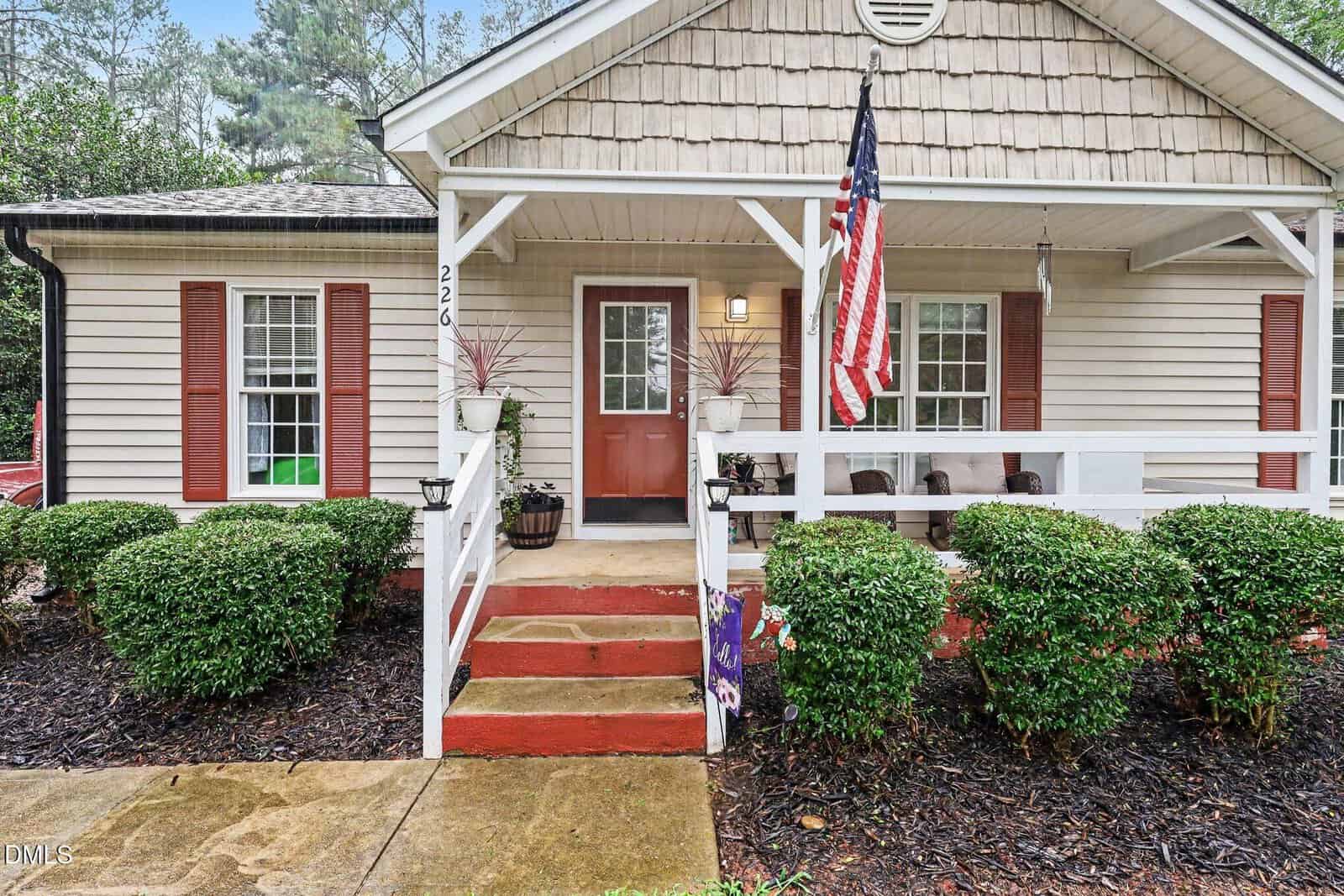 Single-story house with beige siding, red front door, red shutters, white-railed porch, American flag, potted plants, and neatly trimmed bushes. Concrete walkway and steps lead to the porch. Overcast sky and trees in background.