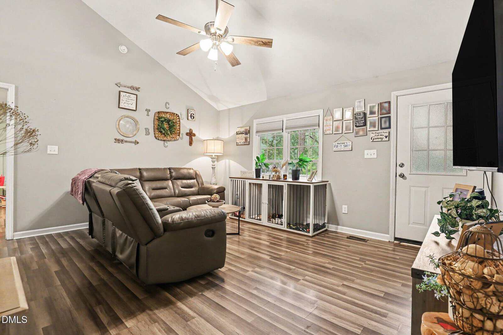 Living room with gray walls, wooden floor, a brown sectional sofa, and a ceiling fan. Decor includes framed pictures and plants. There is a white door and two windows with crate-style storage beneath them.