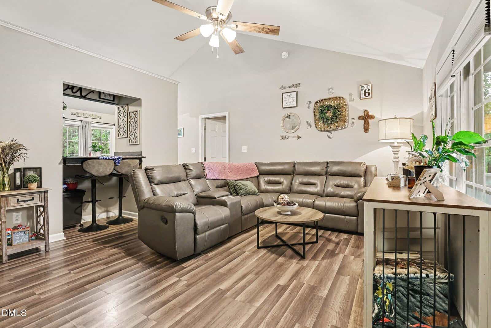 A living room with a gray sectional sofa, round coffee table, ceiling fan, wood floors, and decor on the walls. A window and sideboard with plants are on the right, and a breakfast bar with stools is at the back.