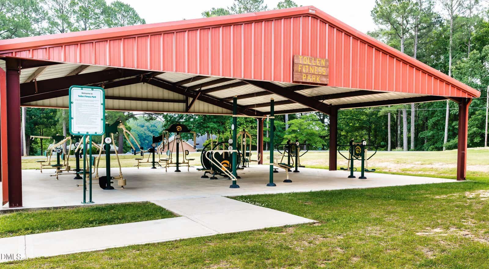 Outdoor gym equipment is arranged under a large red metal pavilion with a sign reading “Woolen Flowers Park.” Trees and grass surround the area, and a playground is visible in the background.