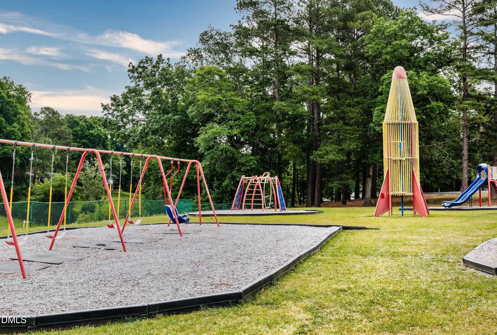 A playground with red swings, a blue slide, a dome climbing structure, and a tall rocket-shaped play tower, surrounded by grass and trees under a partly cloudy sky.