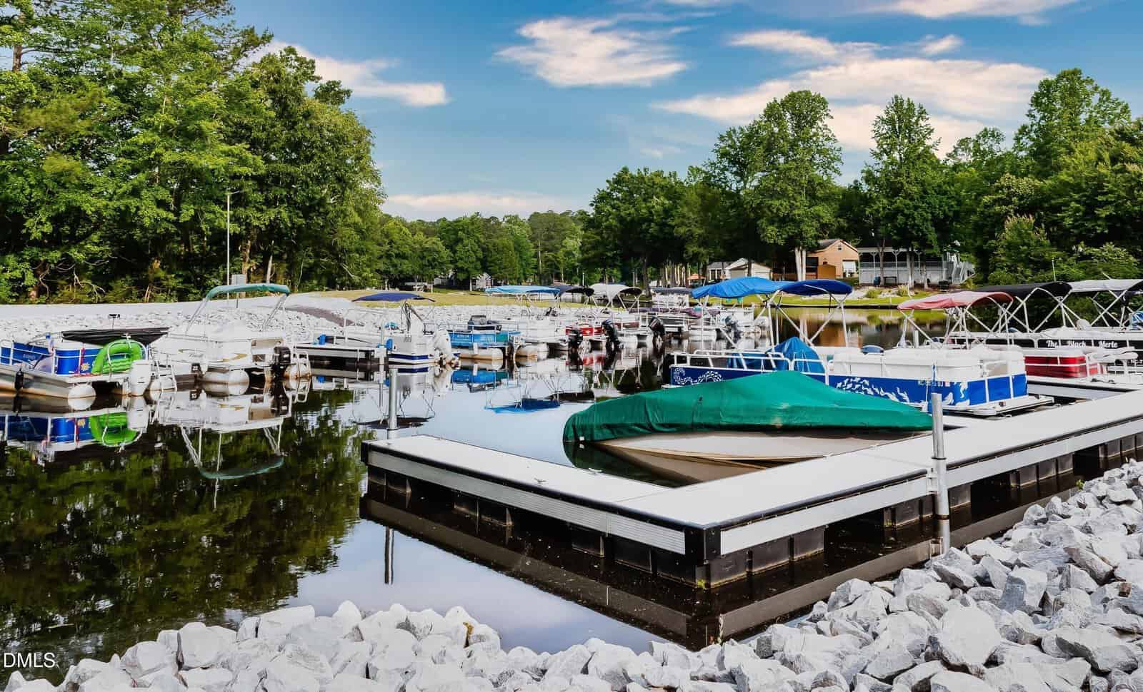 Small boats and pontoons docked at a marina on a calm lake, surrounded by trees and houses; blue sky with scattered clouds reflected in the water.
