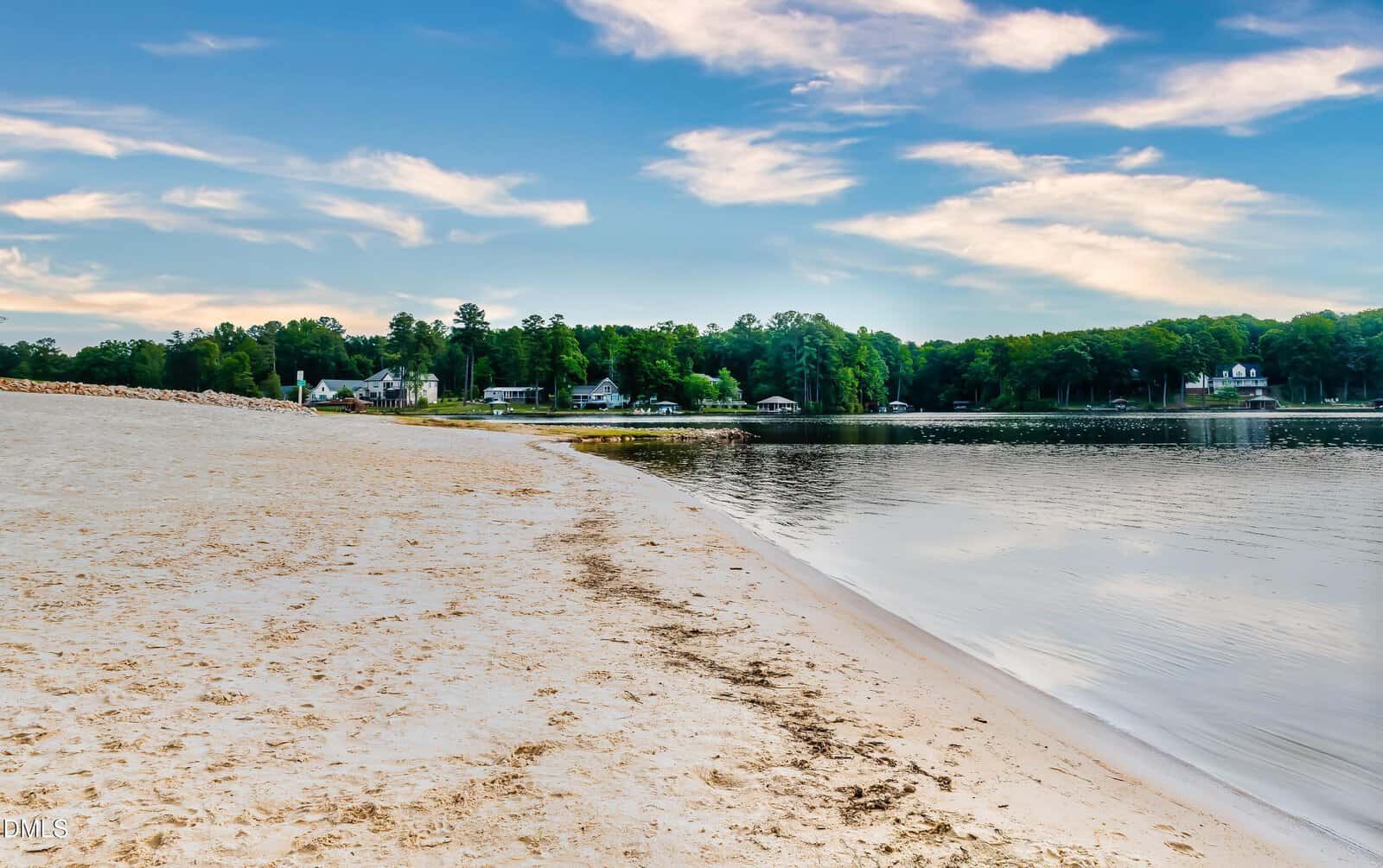 A sandy beach curves along the edge of a calm lake. There are trees and a few houses in the background under a partly cloudy sky. The scene appears peaceful and quiet.